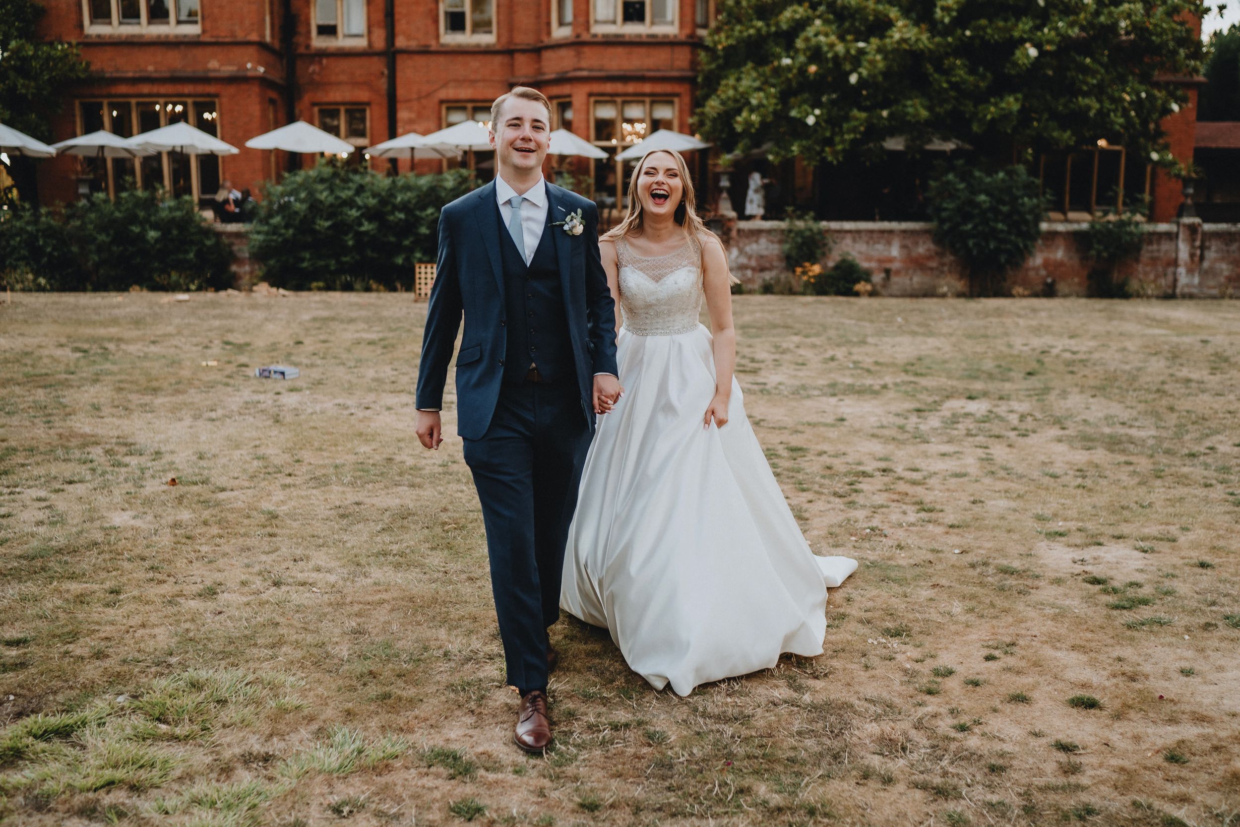 A couple laughs together while walking across a dry lawn in front of a red brick building on their wedding day.
