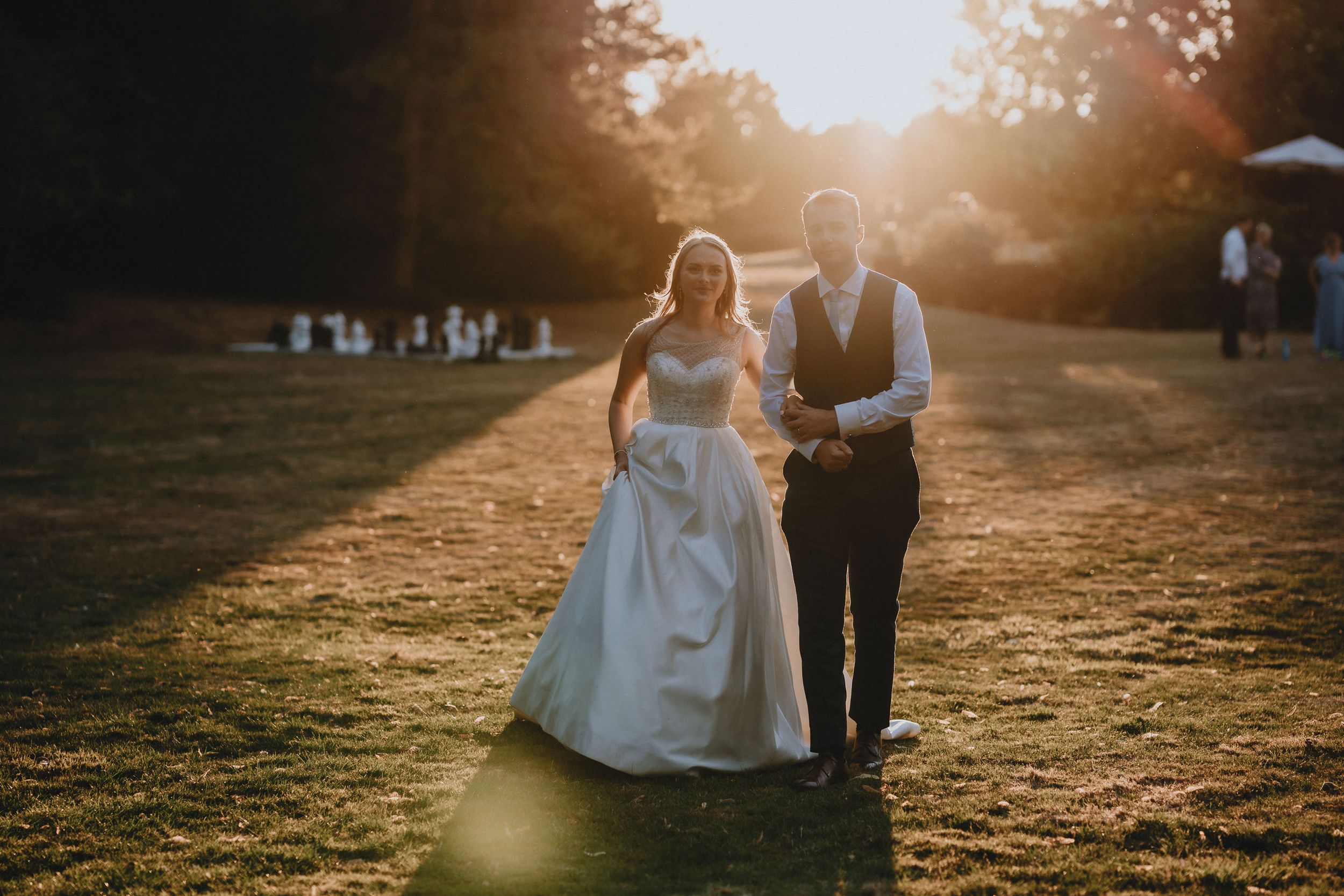 A couple takes a romantic stroll through a sunlit field during golden hour at their outdoor wedding celebration.