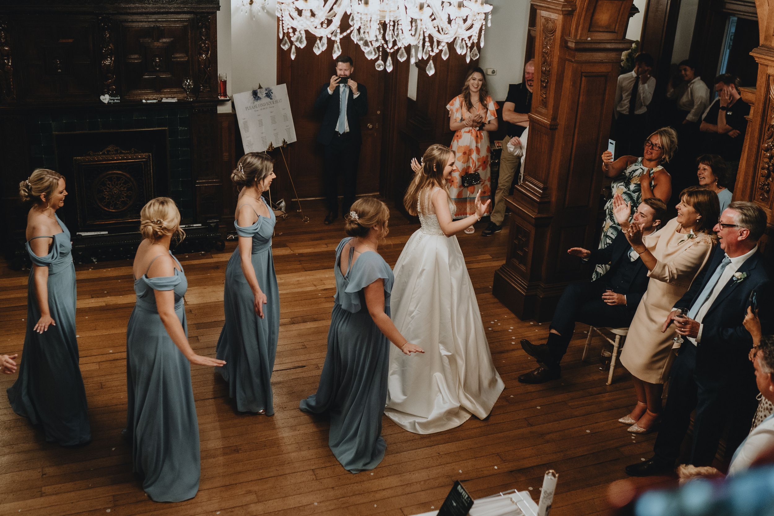 Wedding party stands in formation on wooden dance floor under elegant chandelier during formal ceremony.