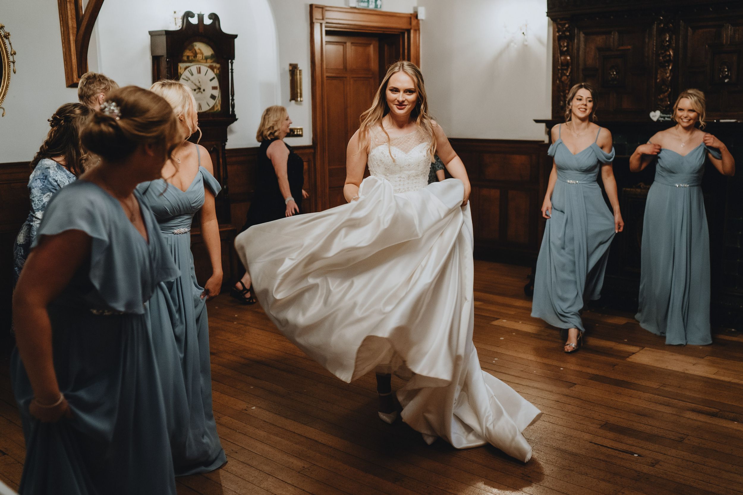 Bridesmaids in flowing blue dresses help a bride with her white wedding gown on a polished hardwood floor.