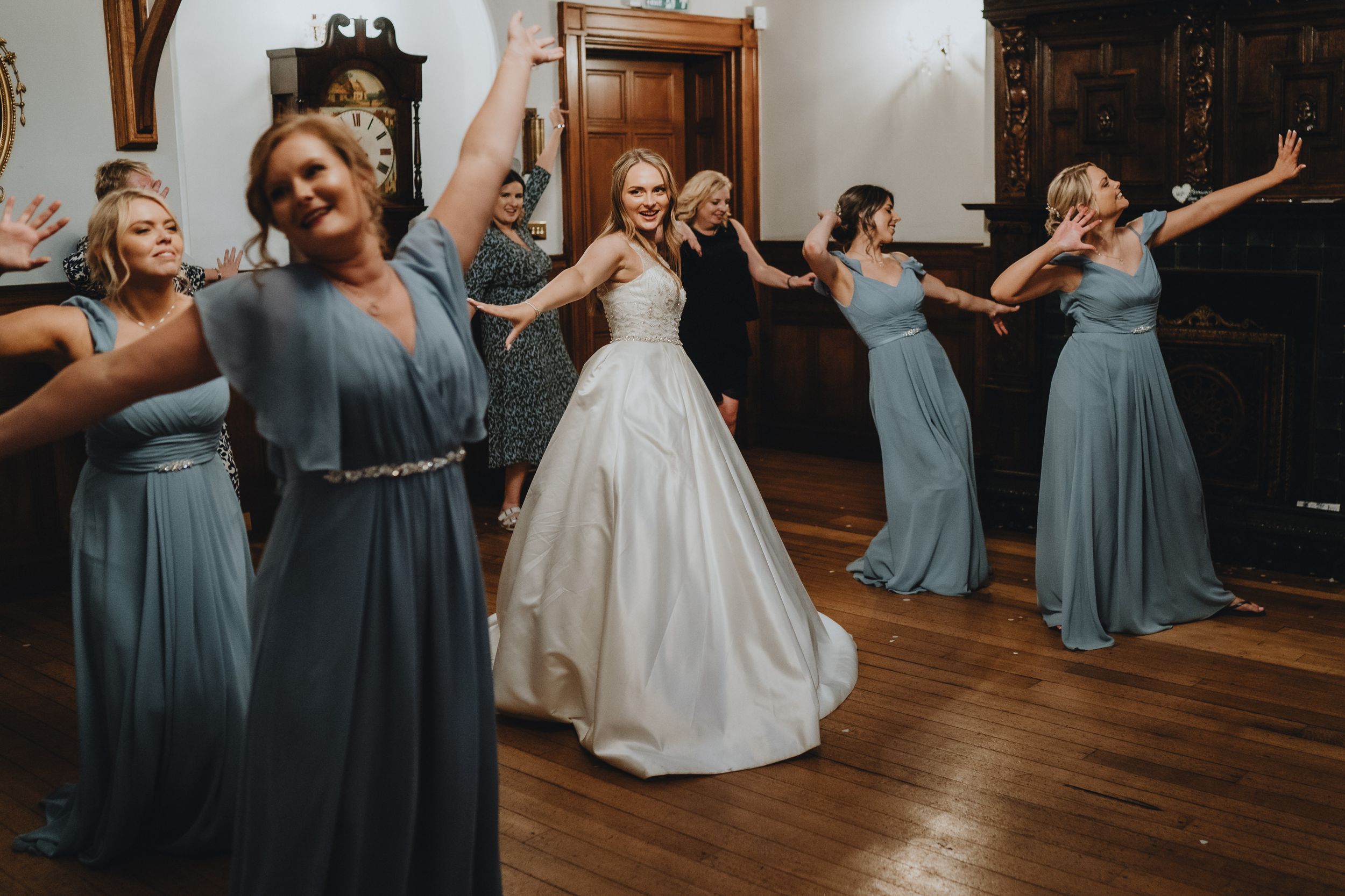 A wedding party dances joyfully in a wood-floored room, with bridesmaids wearing blue gowns surrounding a bride in white.