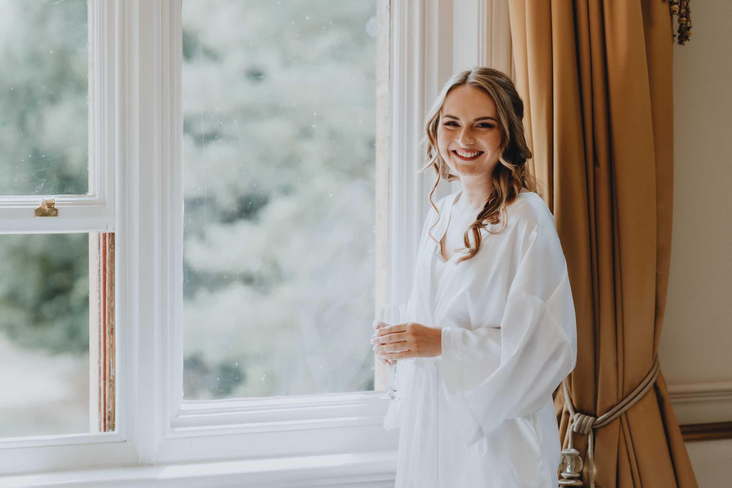 Smiling bride at Holmewood Hall wearing a white robe stands by a window with gold curtains in a bridal prep setting.