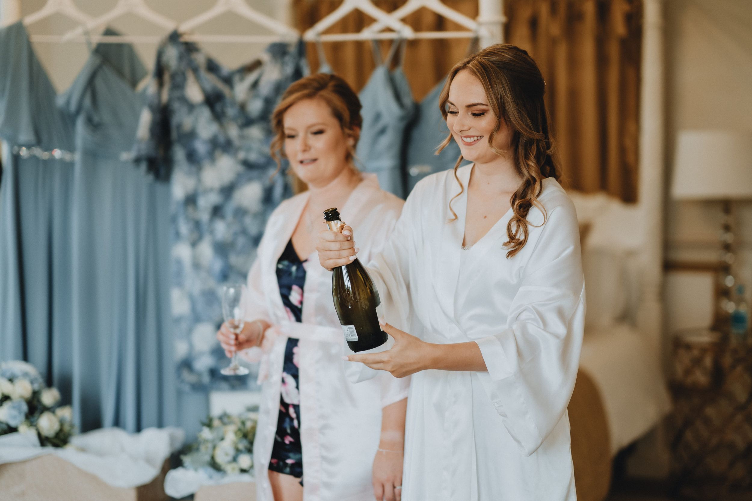 Two people in white robes share a celebratory moment with champagne during wedding preparation.