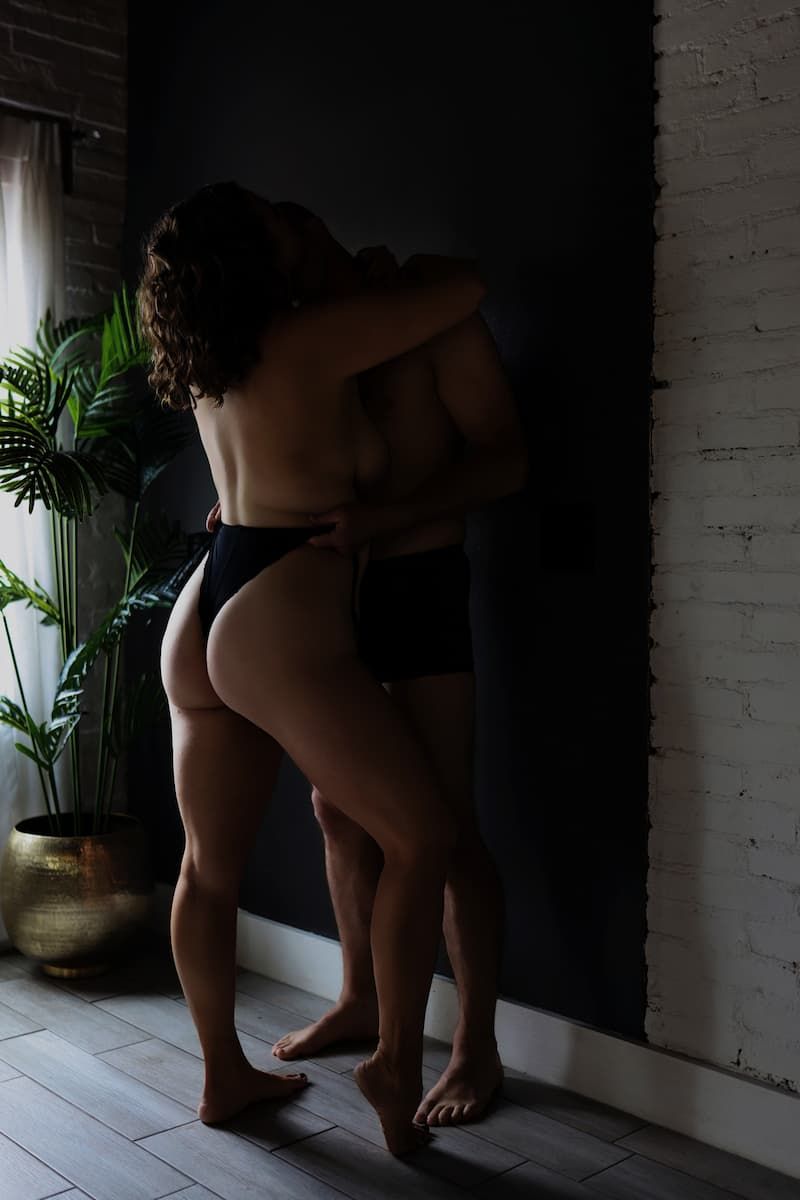Silhouetted figure in dark lingerie stands near a plant against white brick wall in moody lighting.