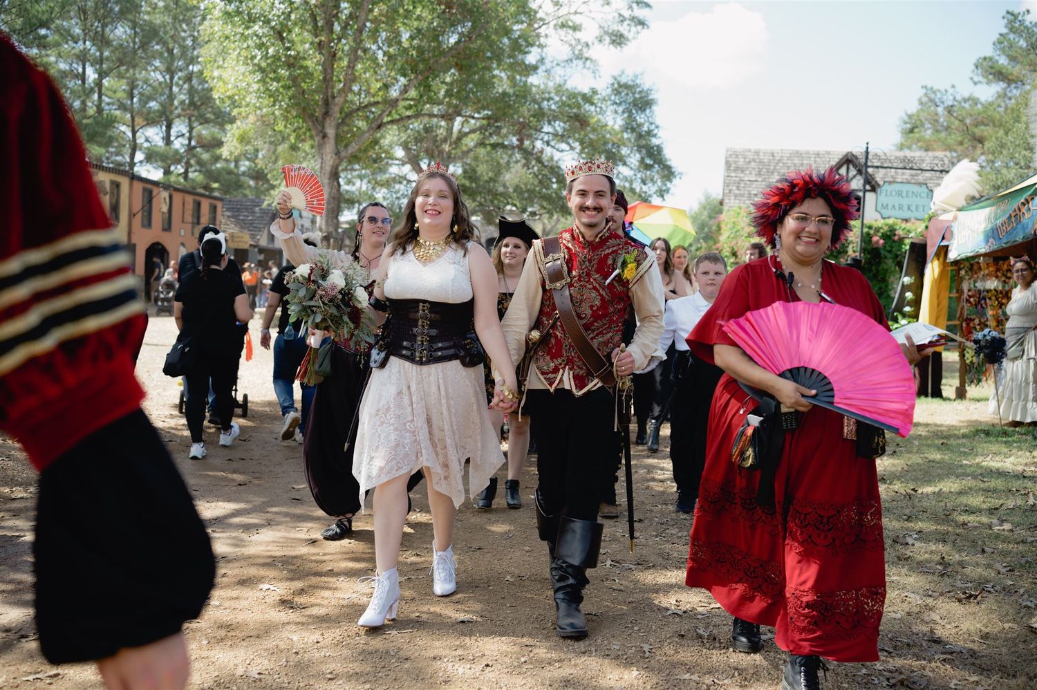 Costumed attendees in period dress walk along a tree-lined path at a Renaissance festival.
