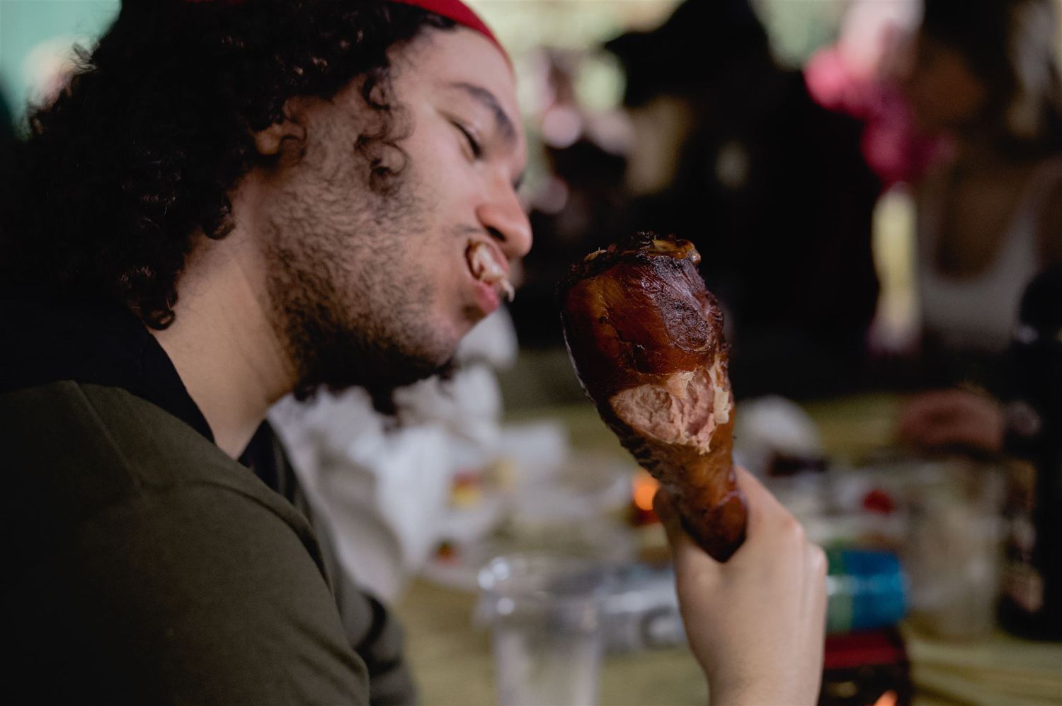 A person with curly dark hair holds up a turkey leg or drumstick while dining at a table.
