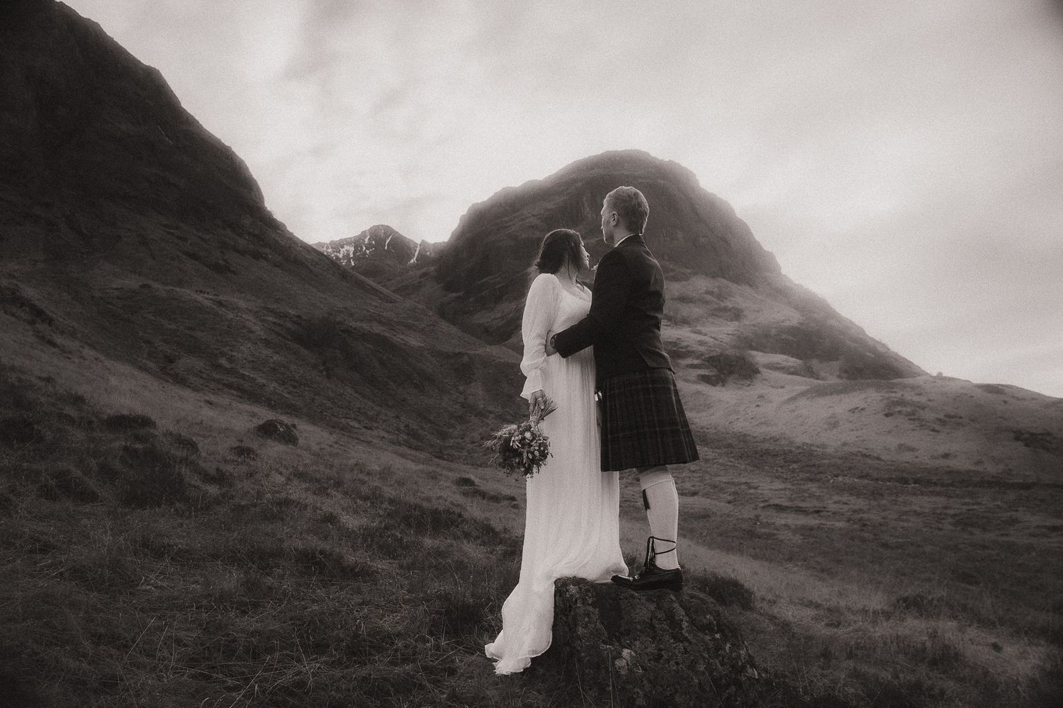 A romantic black and white wedding photo with a couple embracing on a misty Scottish hillside in traditional attire.