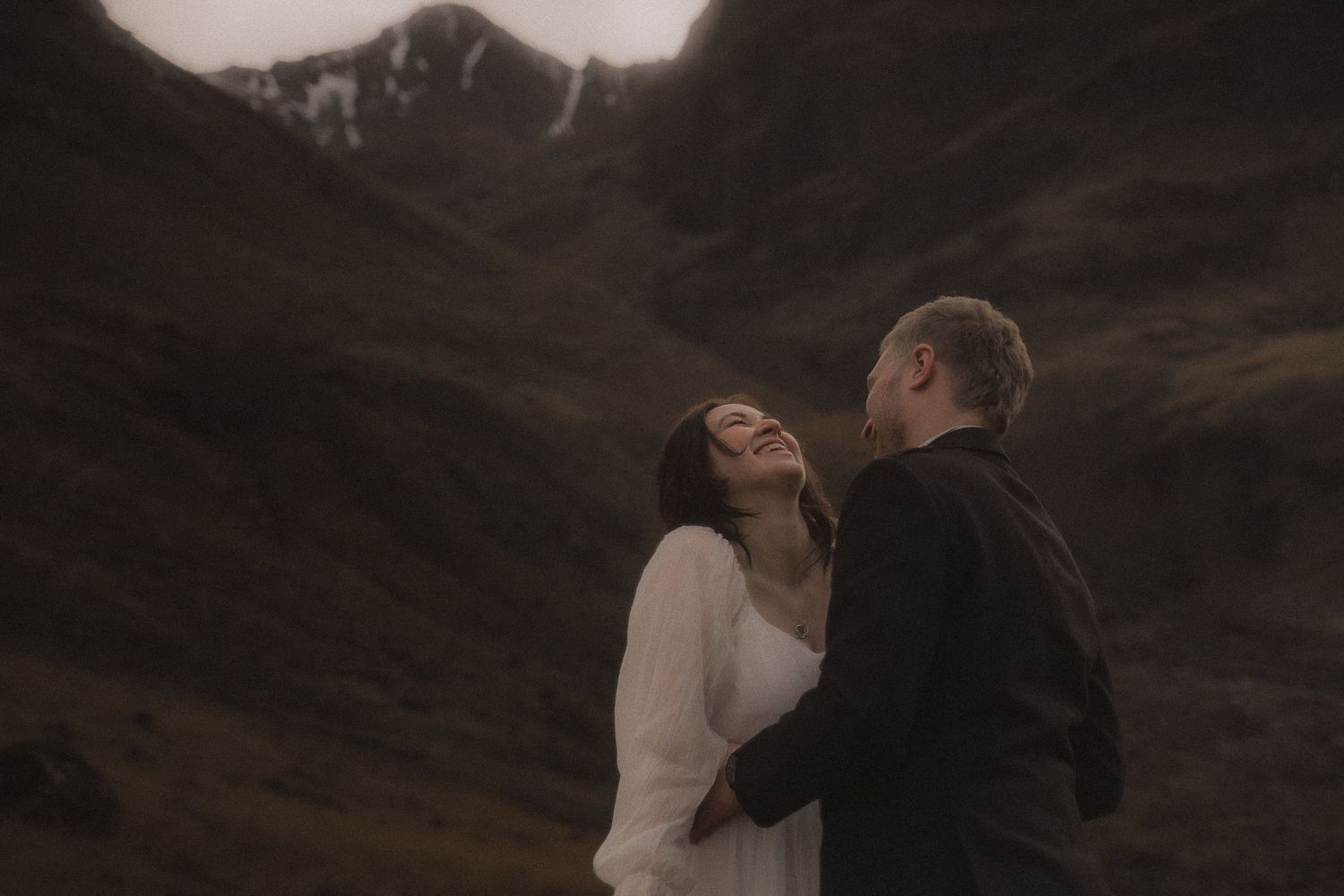 A couple shares an intimate moment of laughter during elopement wedding against a backdrop of misty glencoe mountains in moody lighting.