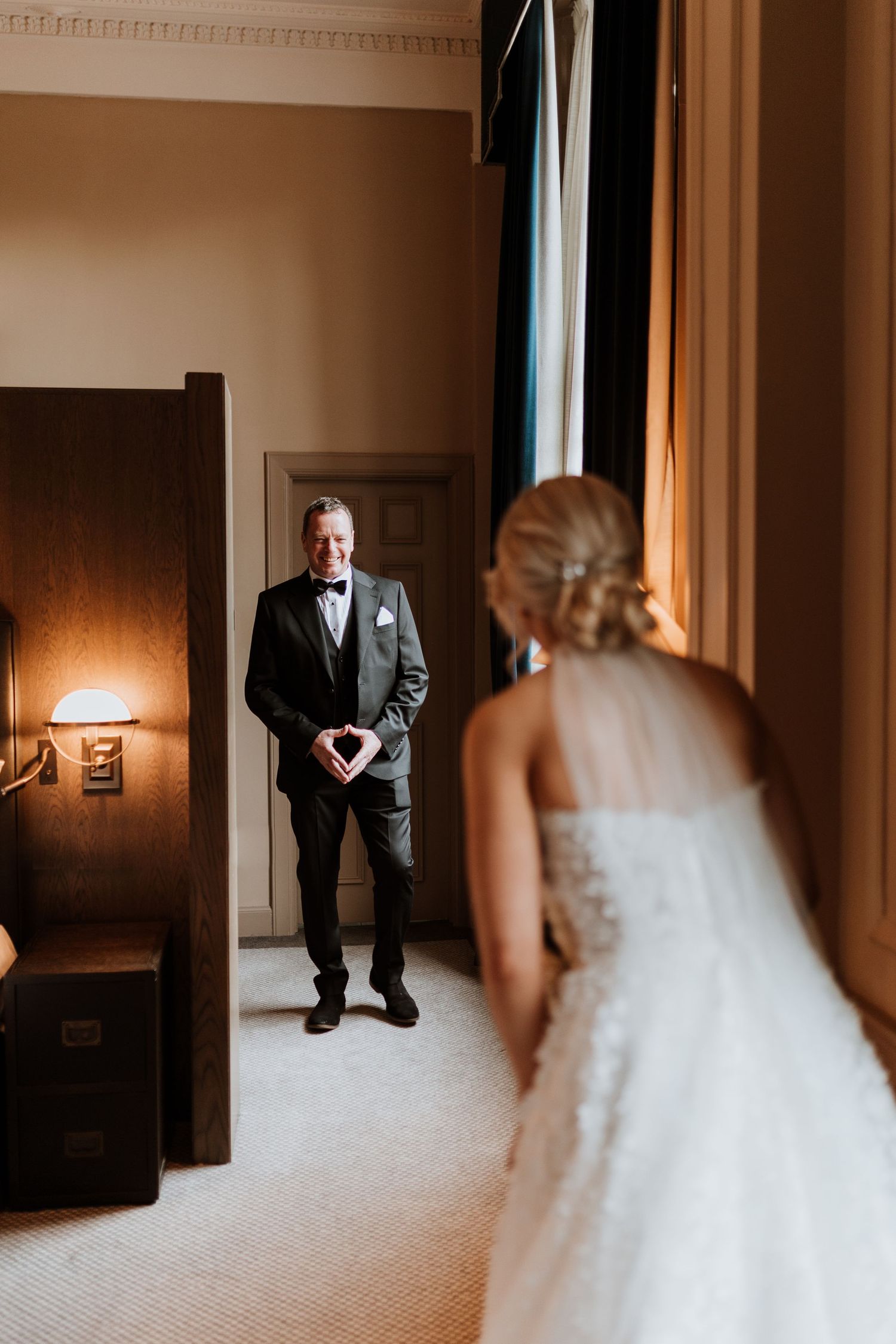 An emotional first look moment between father of the bride wearing a black tuxedo and a bride in her wedding dress in a warmly lit room.