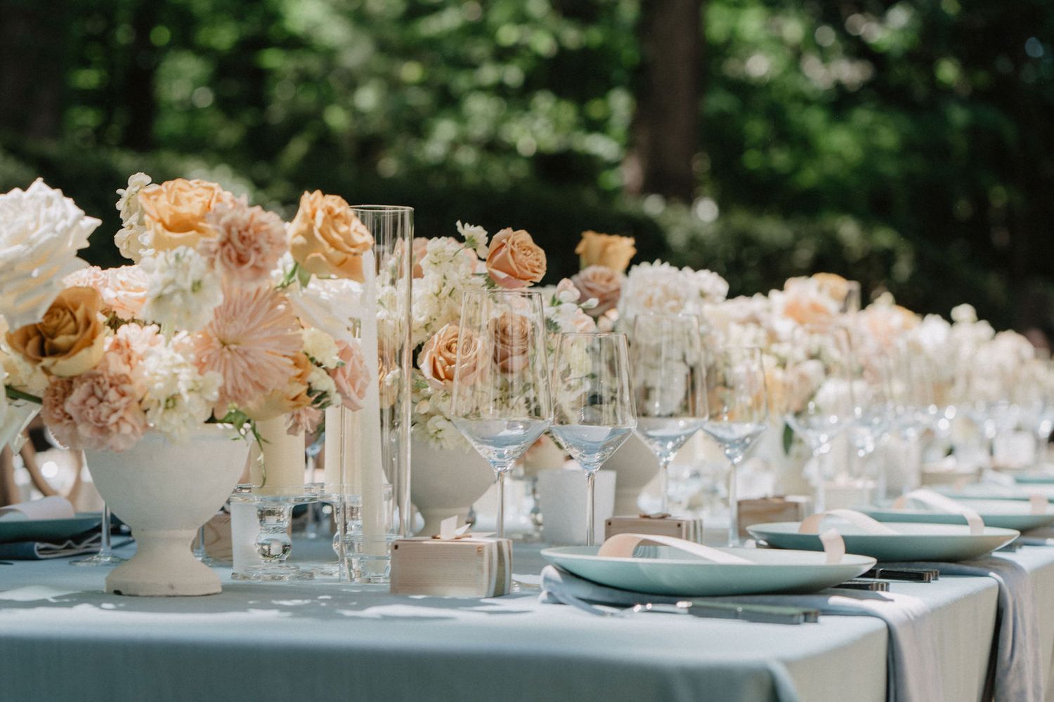 An elegant wedding table setting with peach and cream roses in white vases along a blue tablecloth.