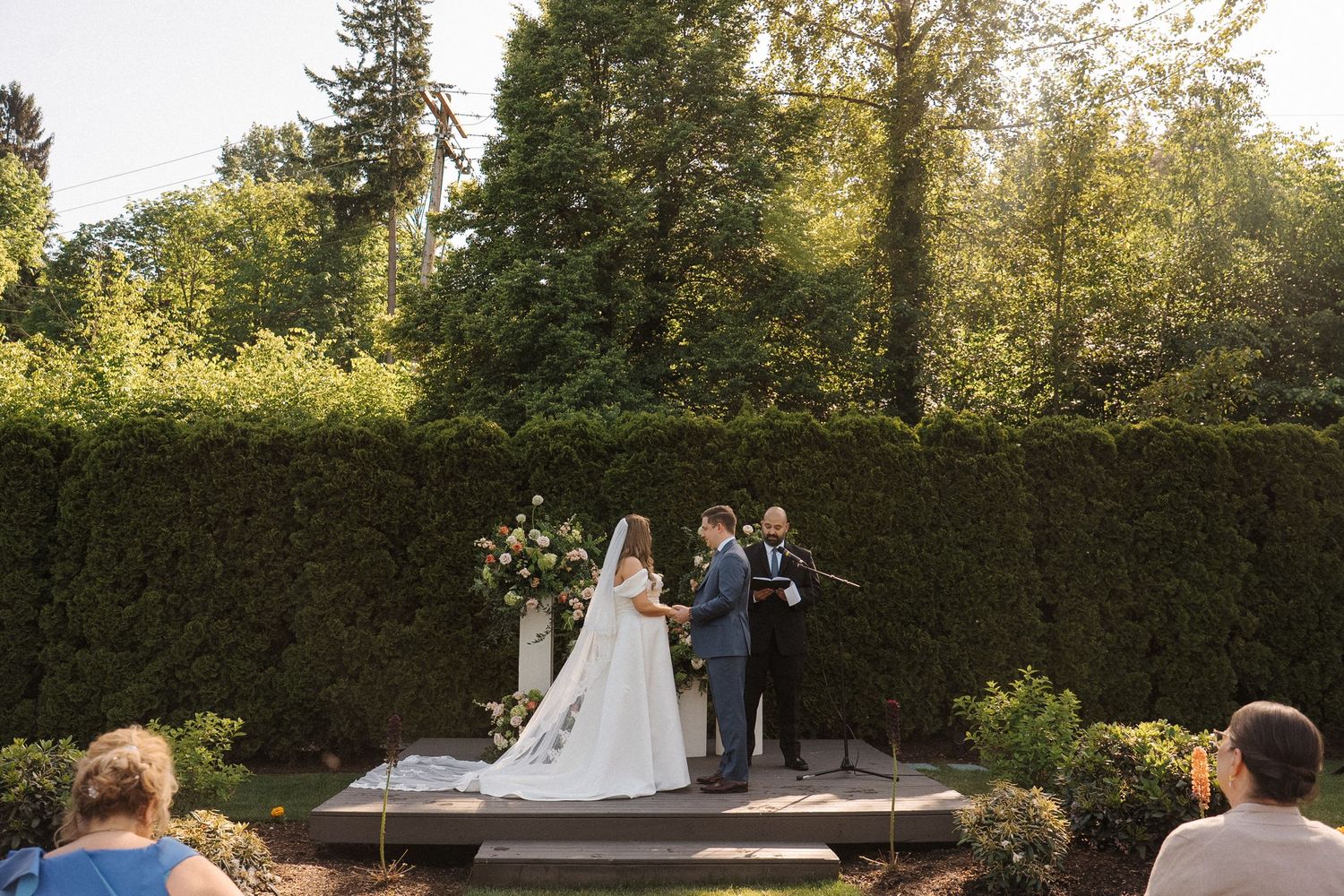 An outdoor wedding ceremony takes place on a wooden platform surrounded by lush green hedges and trees.