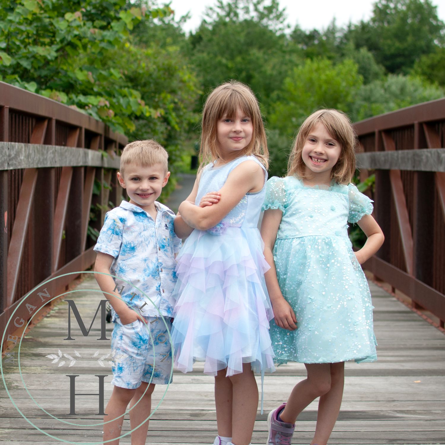 Three kids in summer outfits stand together on a wooden walkway with lush greenery in the background.