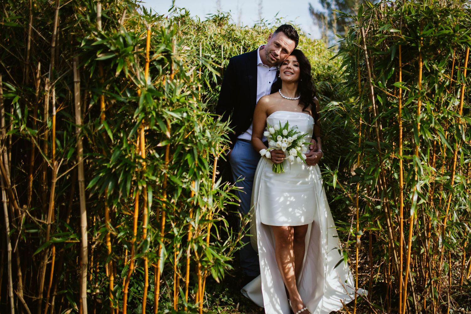 A romantic couple embraces among tall bamboo stalks in a sunlit garden setting.