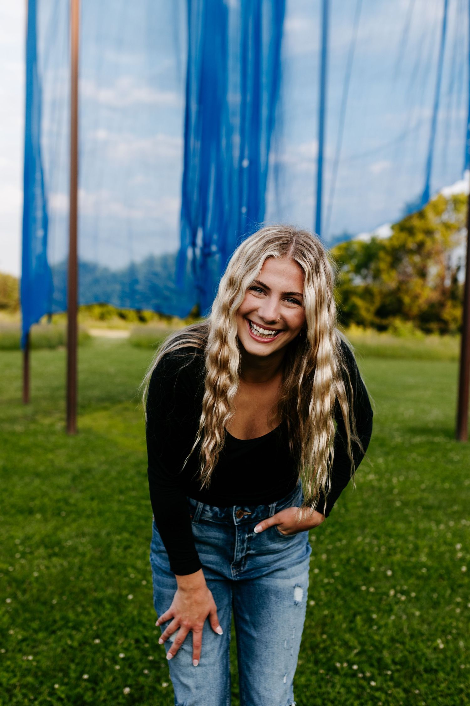 Person with long wavy blonde hair wearing casual attire stands outdoors near tall blue poles on green grass field.