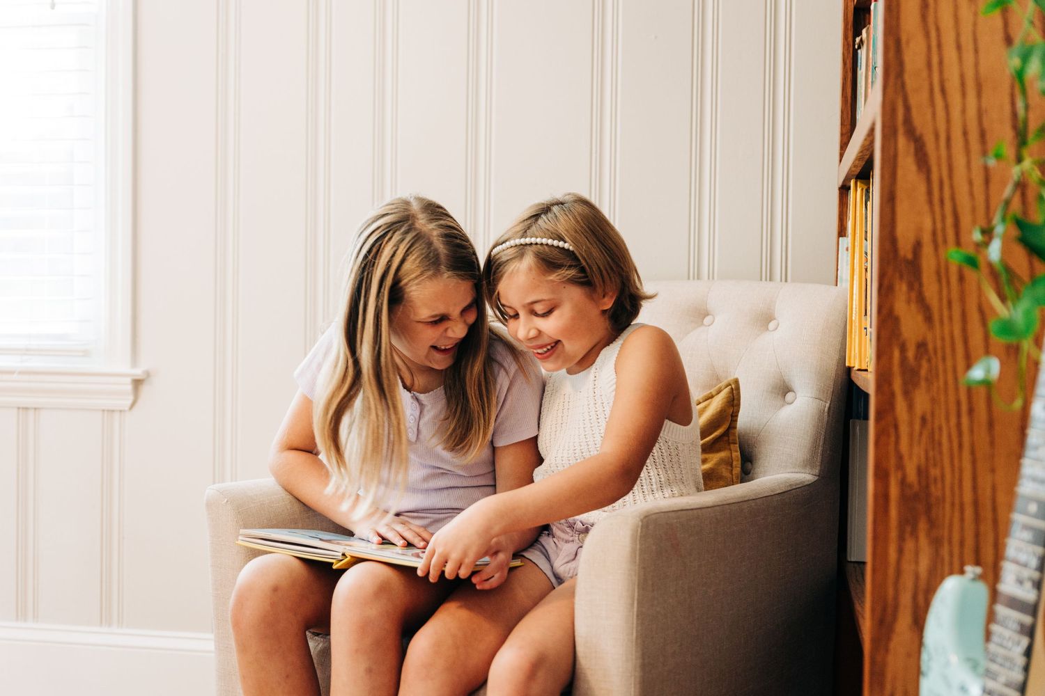 Two sisters sit close together on a couch looking at something in a book.