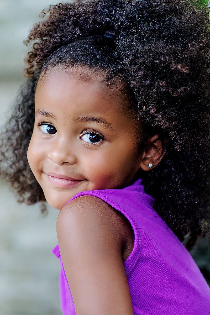 Sweet child in purple top with natural curly hair and bright smile glances playfully at camera in close-up portrait series.