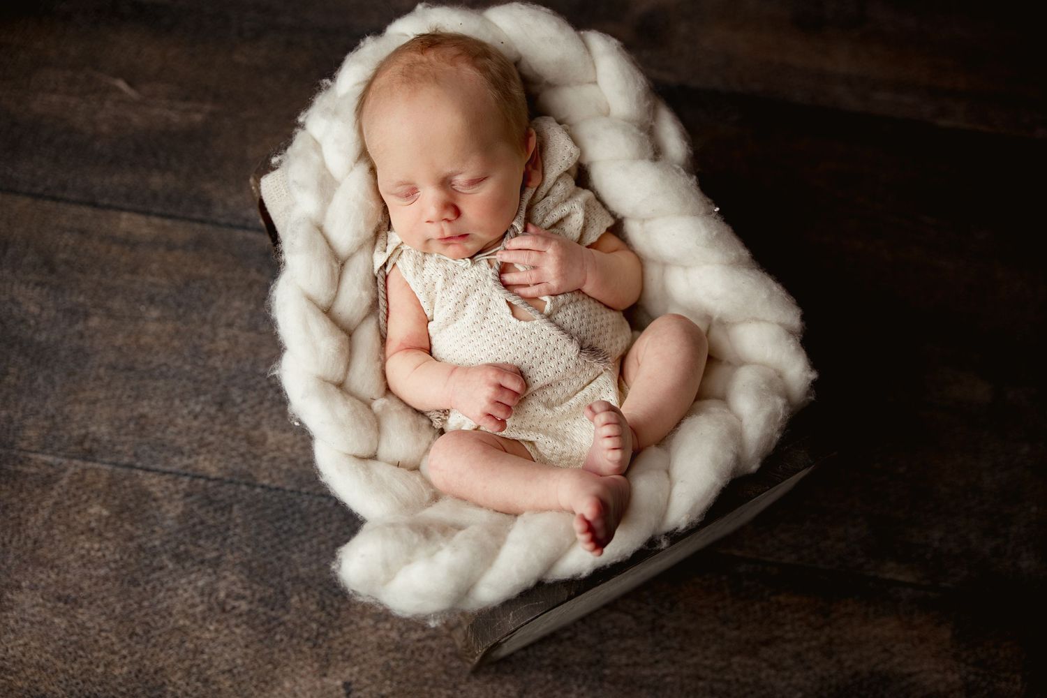 A sleeping newborn wearing white clothing nestles peacefully in a cozy cream-colored wool blanket on dark wooden flooring.