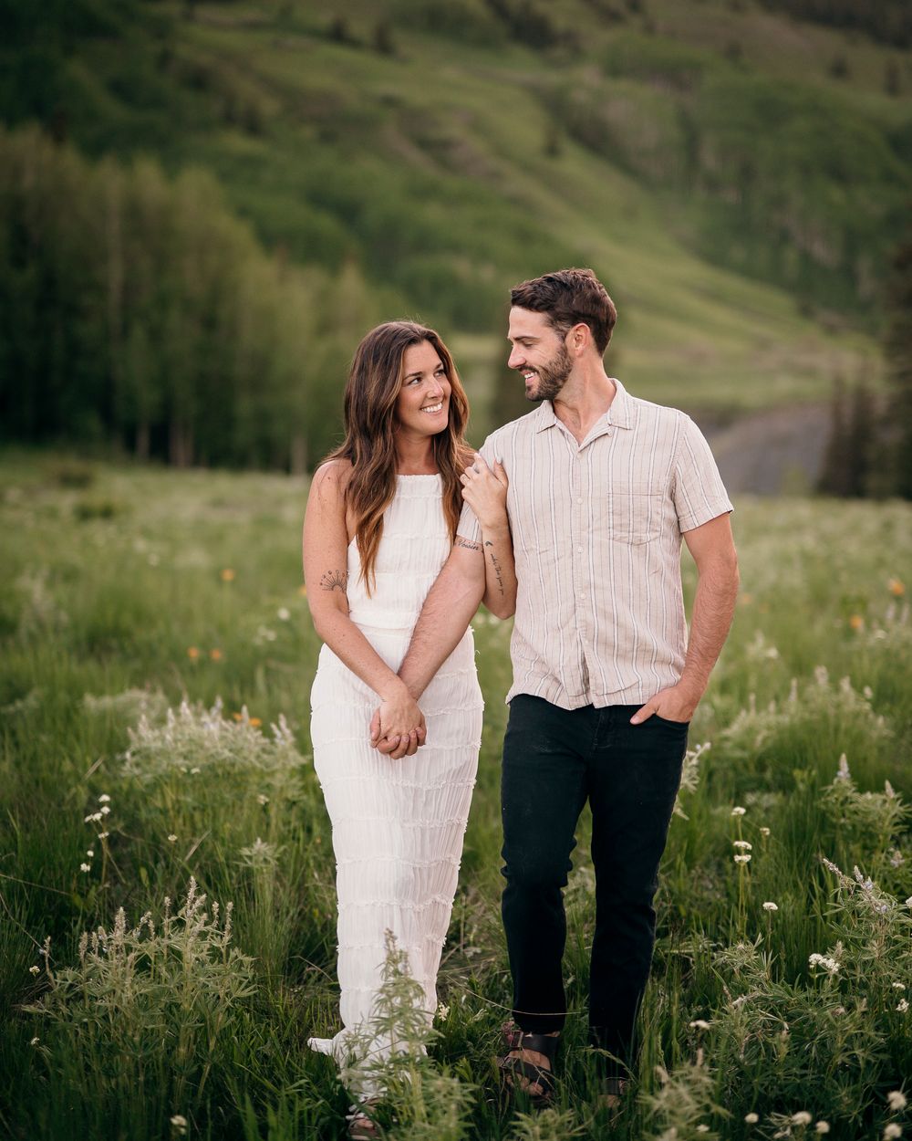 Romantic couple standing close in a mountain meadow wearing light summer outfits.