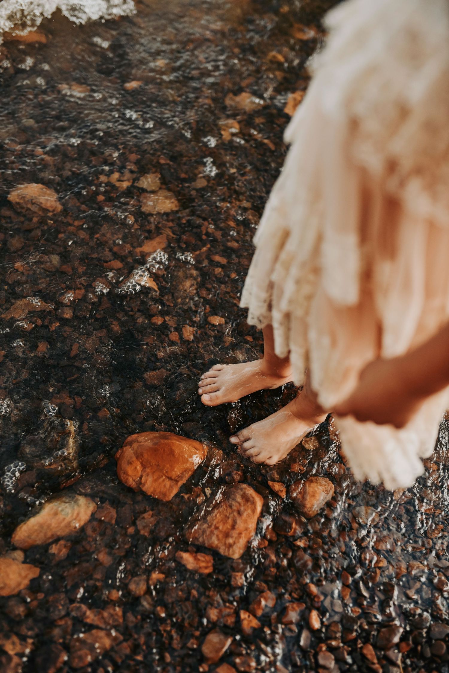 during her family photo a little girl in a lace gown stands barefoot in the water on the edge the lake as the waves splash her toes