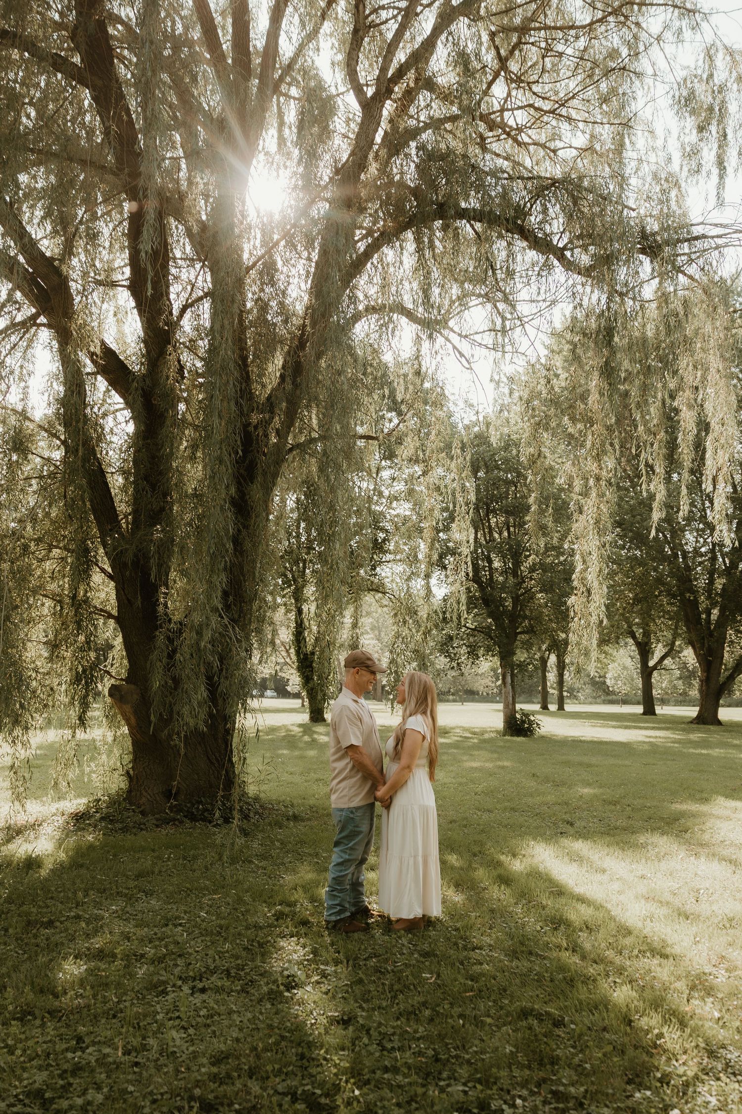 A couple holds hands while walking through a serene park with willow trees on a sunny afternoon.