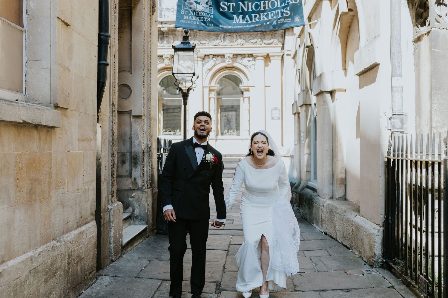 A couple in formal wedding attire walks together through a historic stone archway in Bath, England.