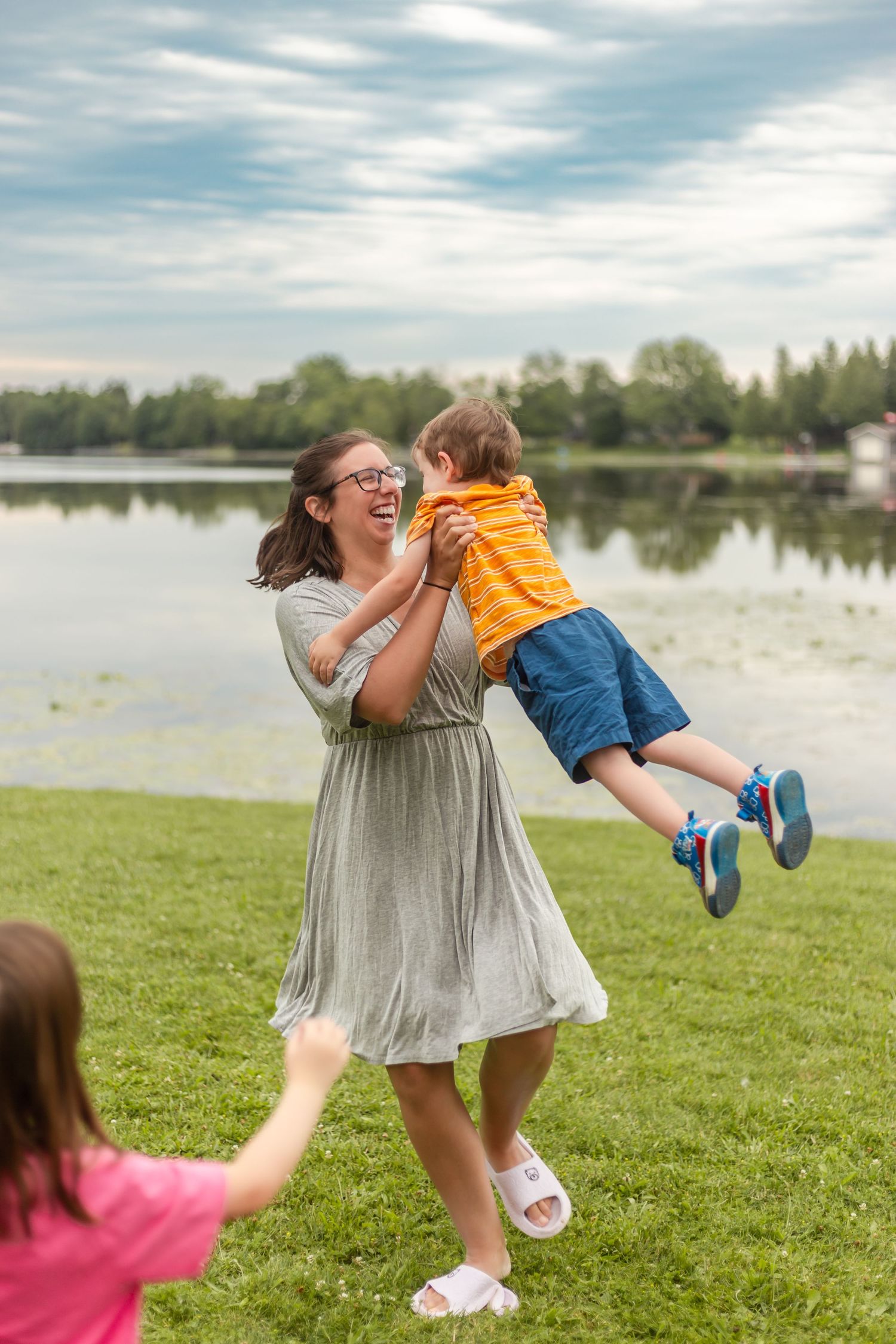 Joyful moment at a lakeside park as a child in orange shirt is lifted playfully into the air on a sunny day.