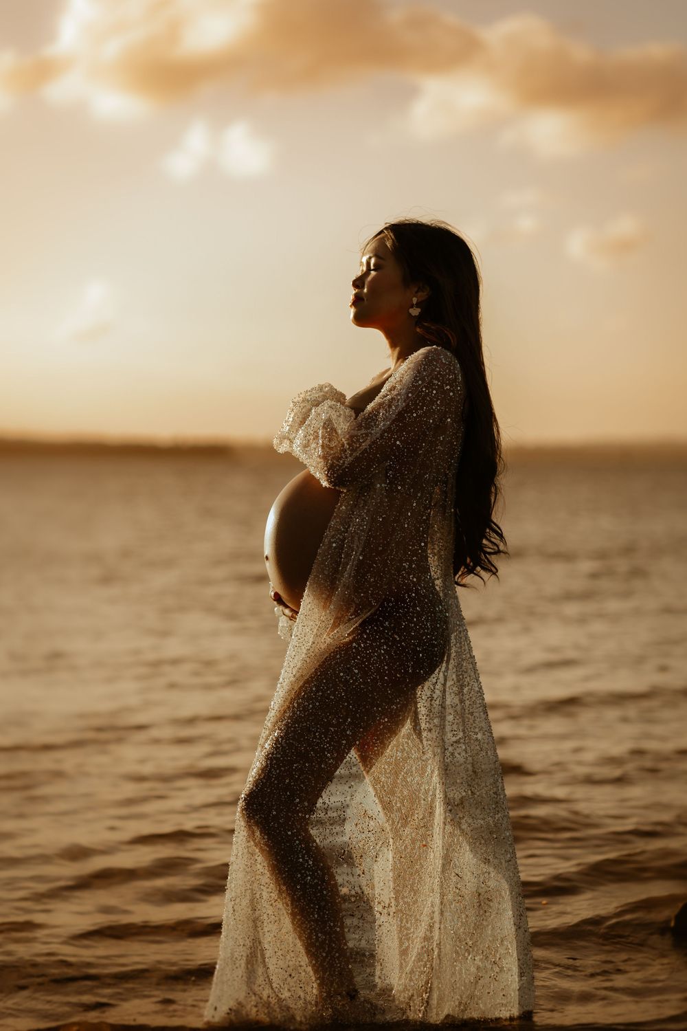 A silhouetted pregnant figure in a flowing dress stands in ocean waters during golden sunset.