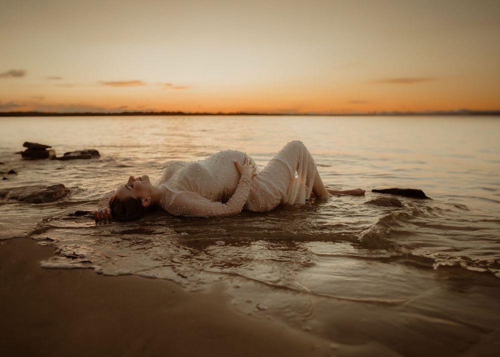 A figure in white dress lies on wet sand at the beach during golden sunset with calm waters and rocks in view.