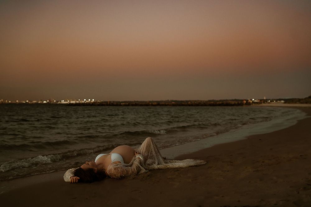 A person lies on a sandy beach at sunset with city lights twinkling along the distant shoreline.