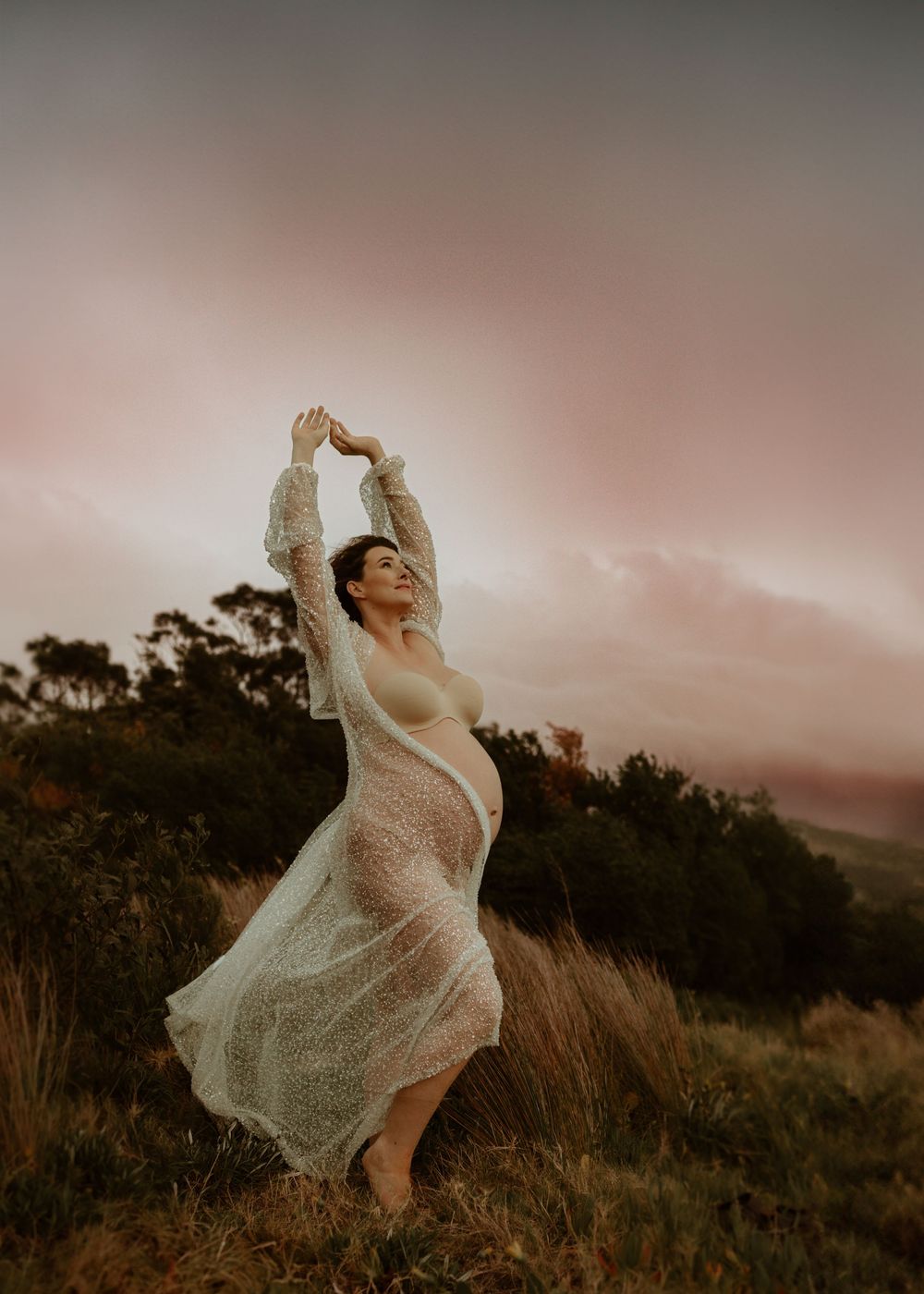 Silhouette wearing sheer white dress dances gracefully in field at sunset with mountains in background.