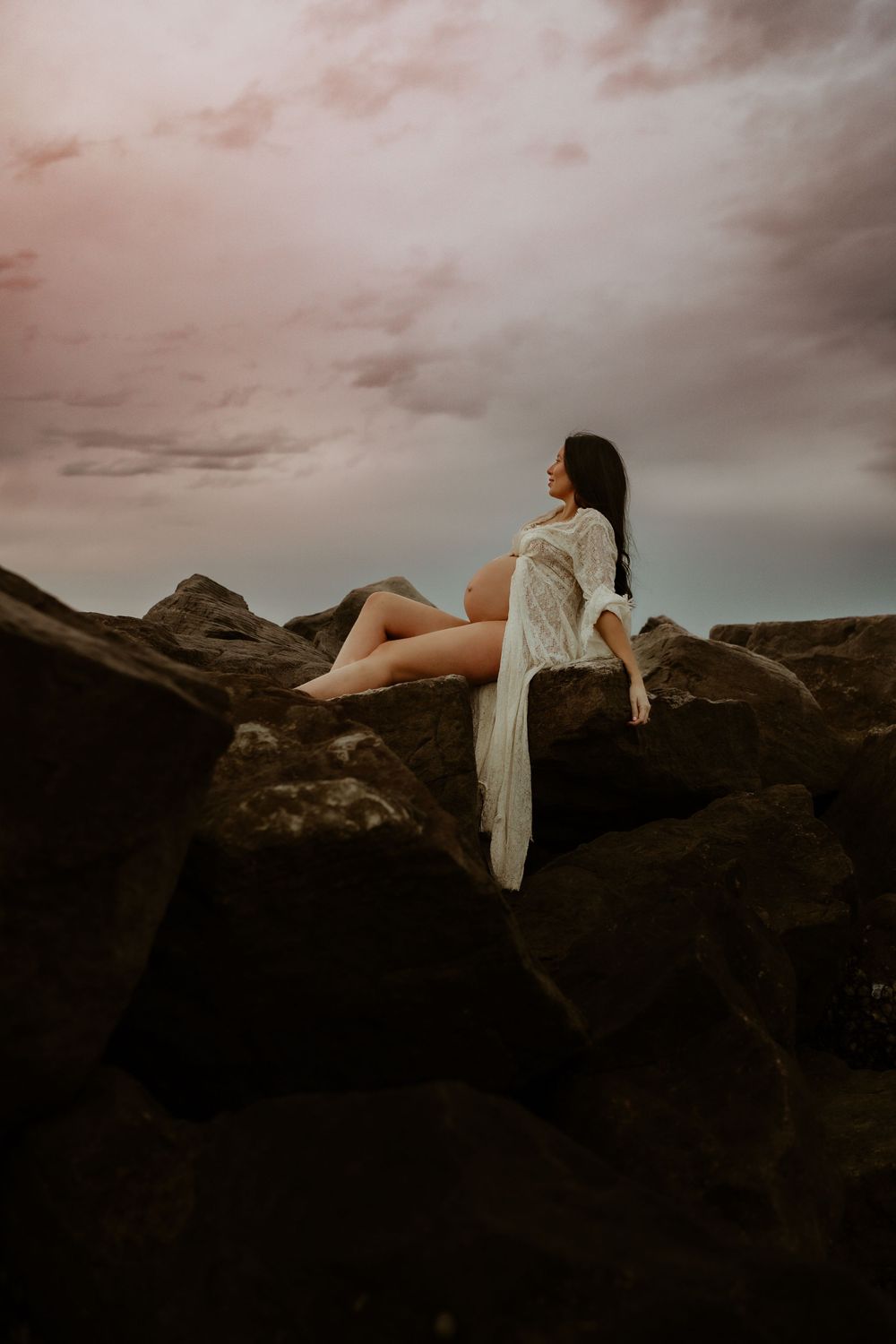A person in a flowy white dress sits on dark rocky terrain against moody pink-tinted clouds at sunset.