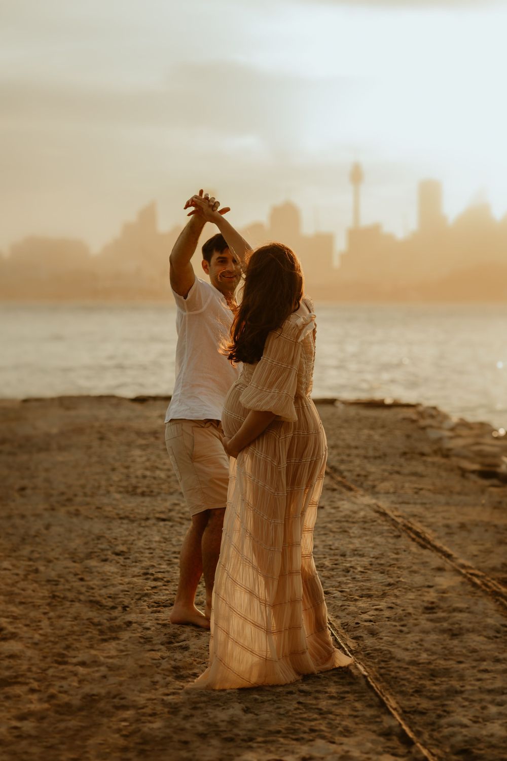 Romantic couple dancing on beach at golden hour with city skyline silhouette in background.