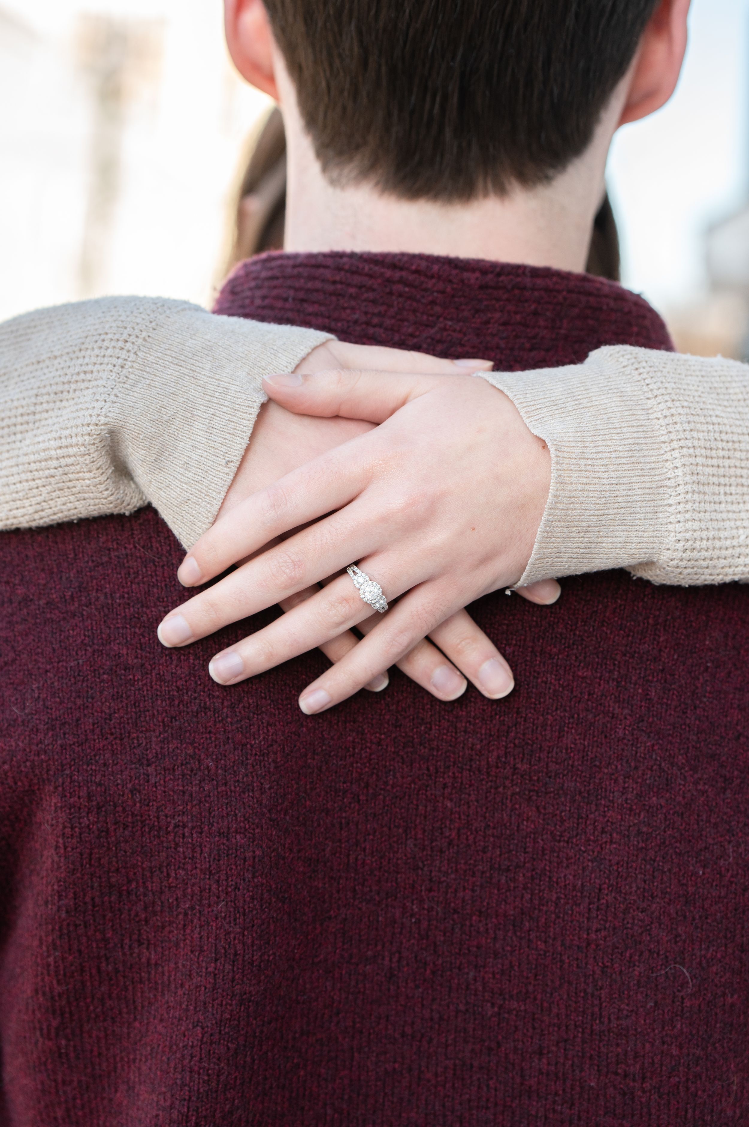 Close up of hands embracing from behind showing an engagement ring on a finger against burgundy and beige sweaters.