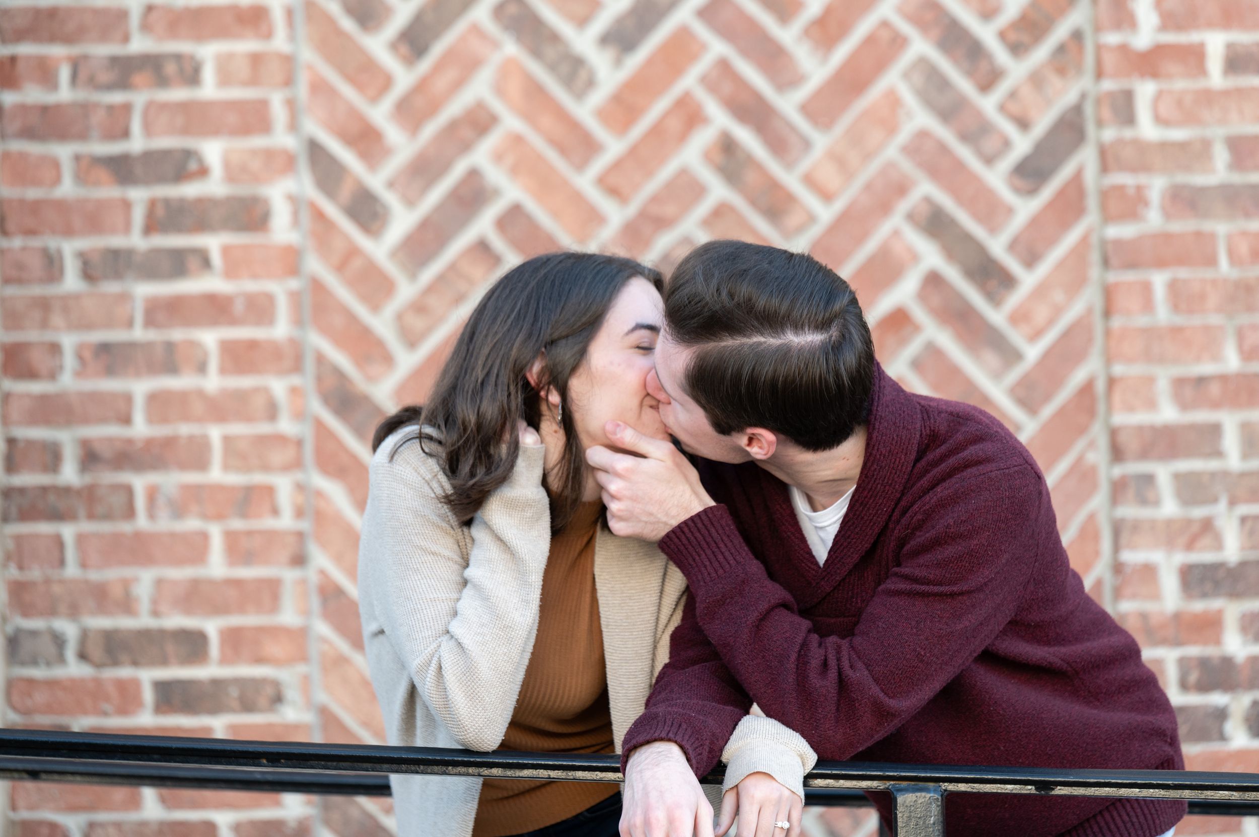 Two people lean in close to whisper secrets while sitting together on a bench against a brick wall.