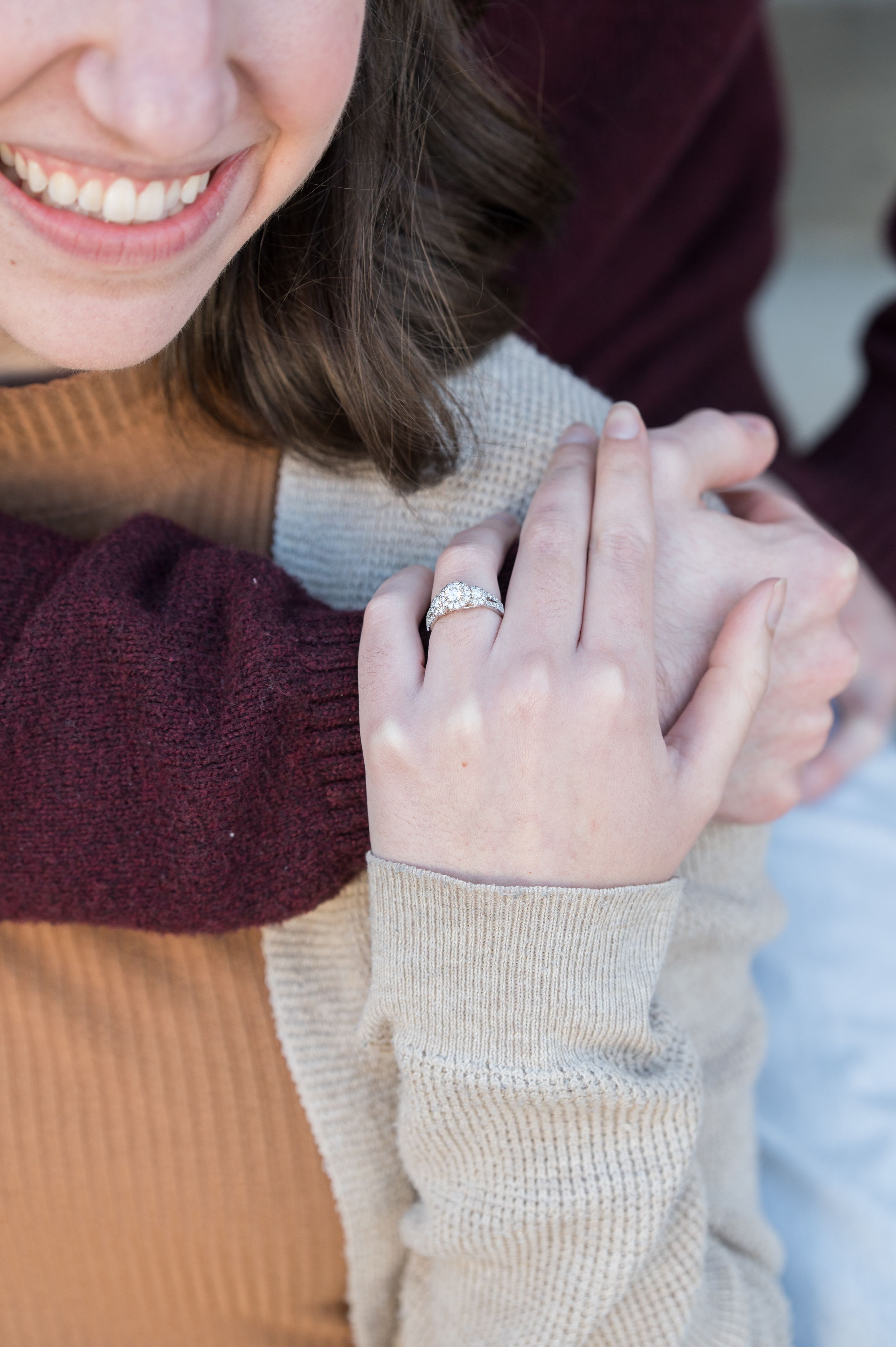 Close-up of an engagement ring being shown on a hand with burgundy and tan sweater sleeves visible.