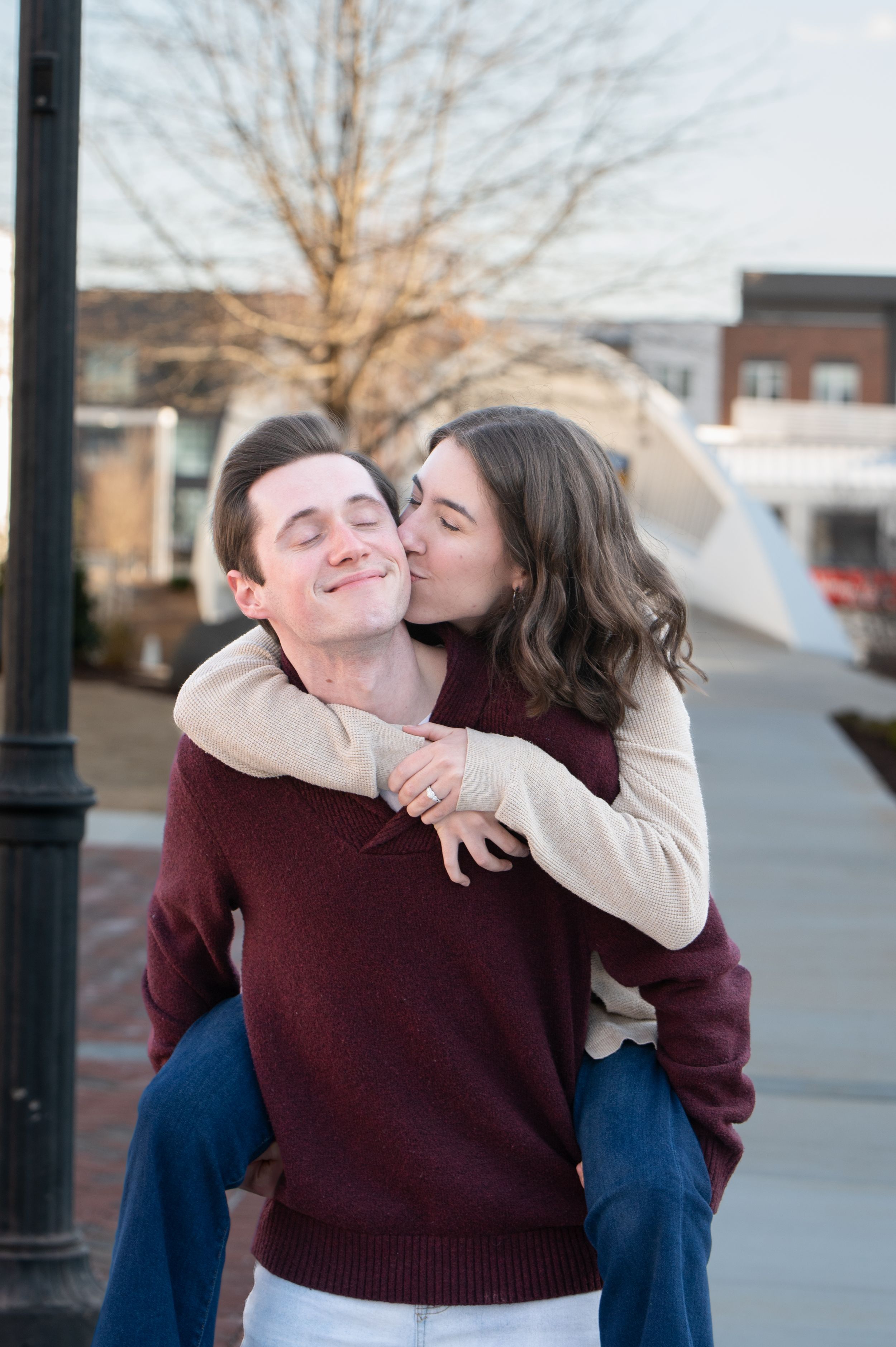 A couple shares a playful piggyback moment on a city sidewalk, wearing matching burgundy and beige sweaters.