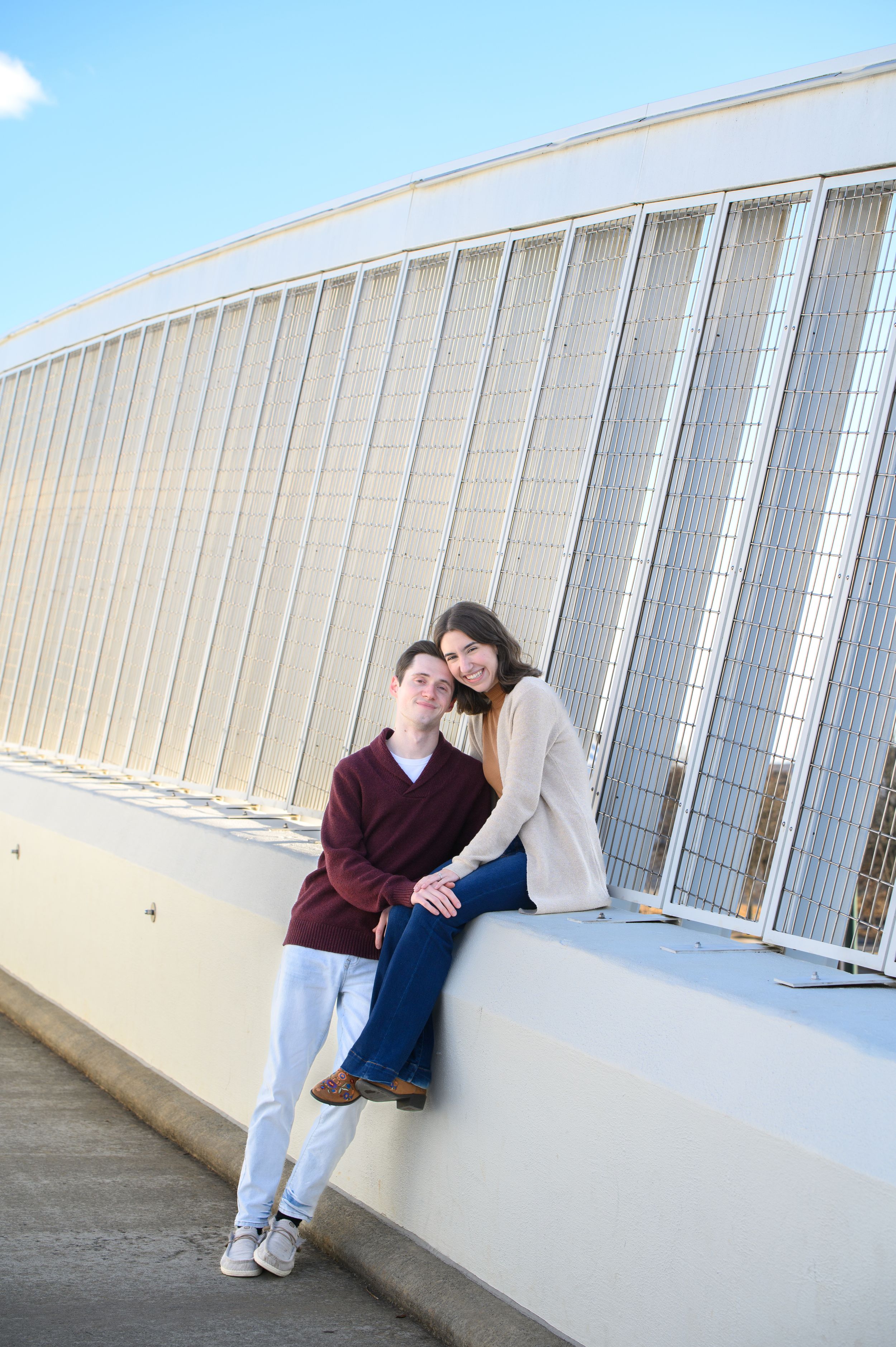A couple poses together on a white ledge against an industrial metal wall on a sunny day.
