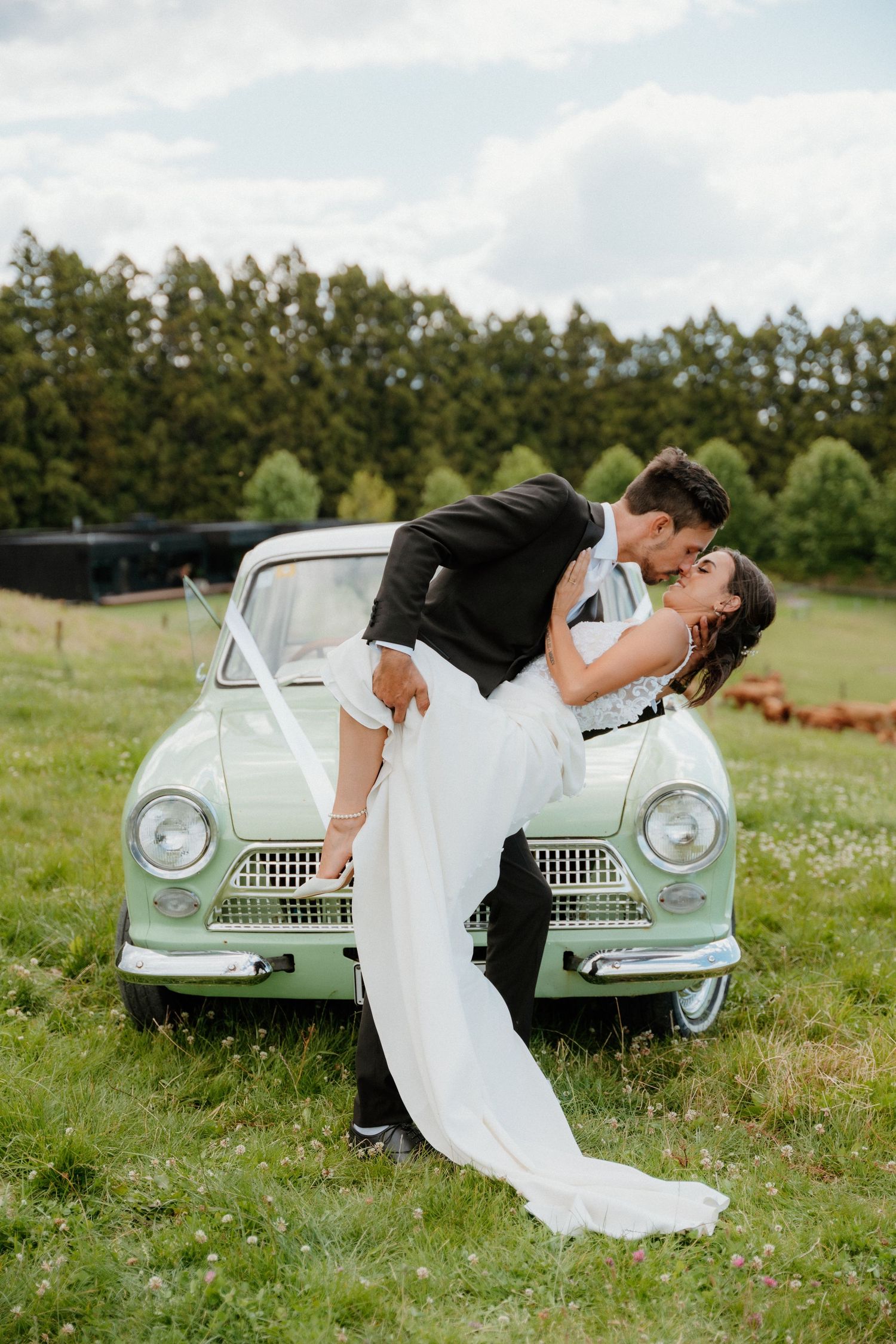 A newlywed couple shares a romantic dip kiss in front of a mint green vintage car in a grassy meadow.