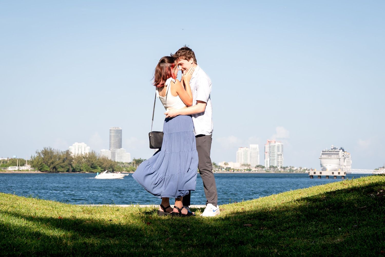 Sunset proposal on a Miami dock with the skyline in the background and the groom kneeling as she reacts with emotion.