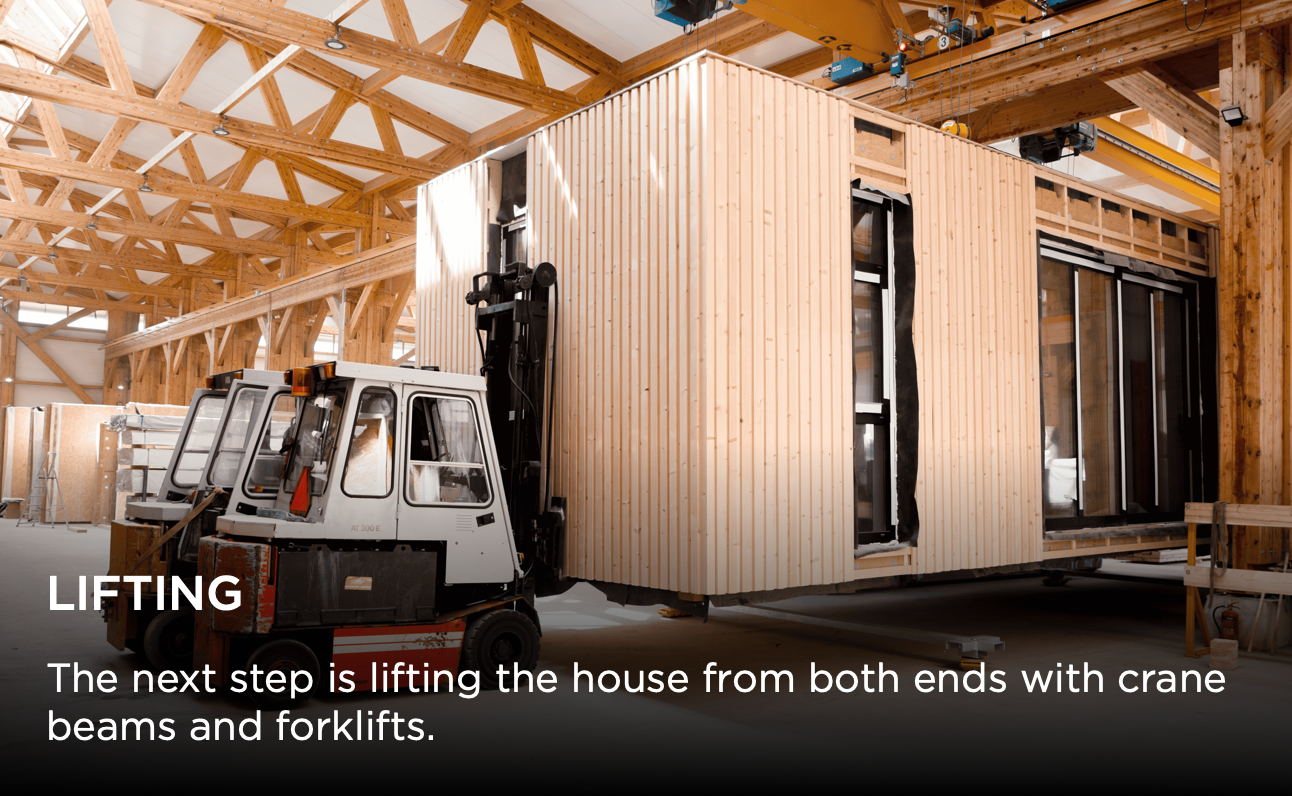 Construction workers use crane beams and forklifts to lift a modular house section inside a warehouse facility.