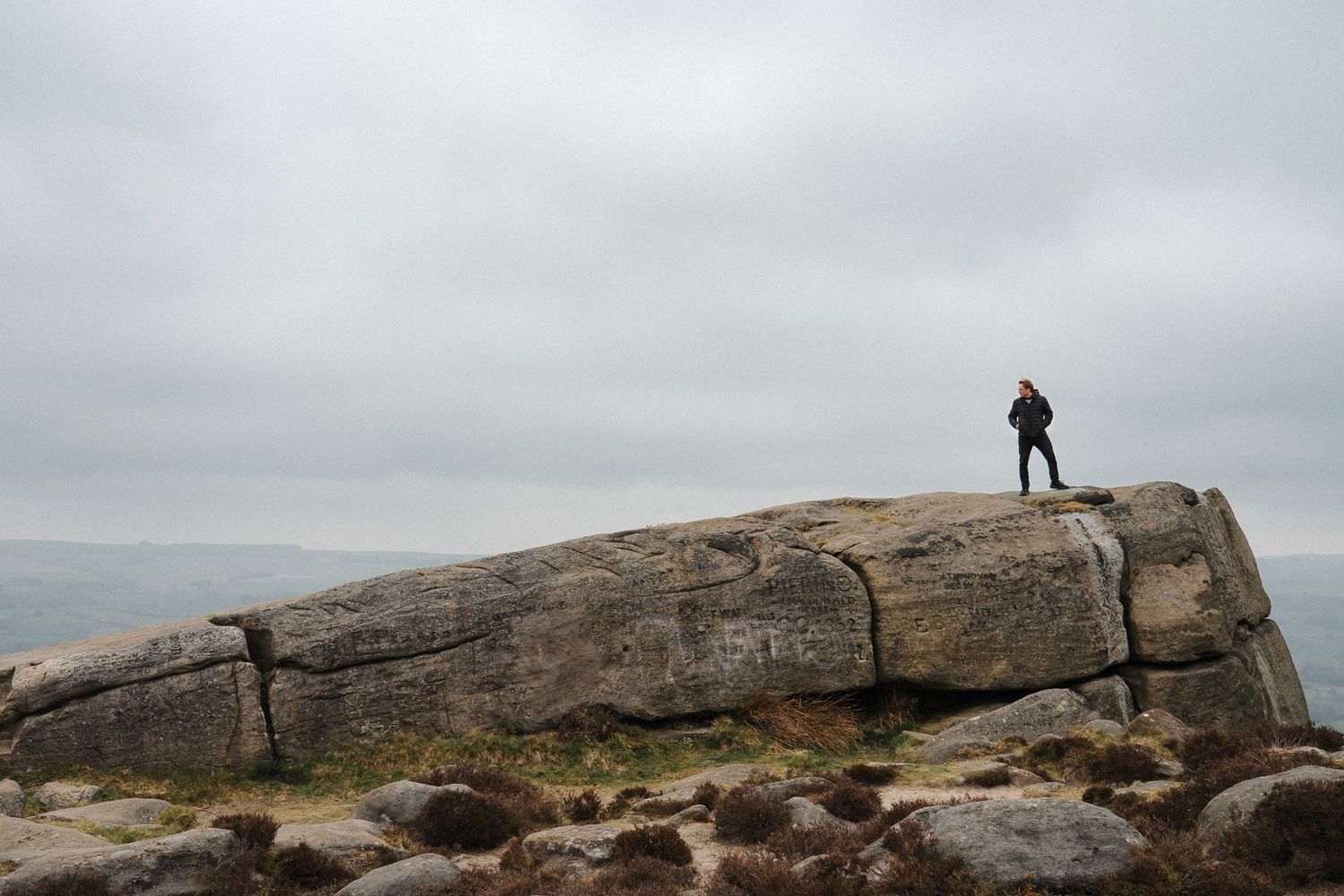 A silhouetted figure stands atop a large rocky outcrop overlooking a misty coastal landscape on an overcast day.