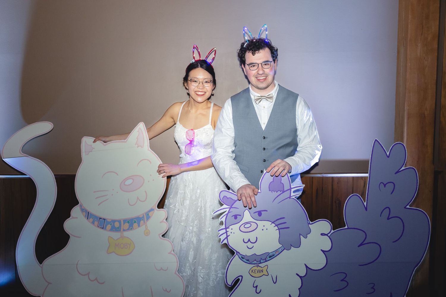 A couple poses in front of playful cat-themed cardboard cutouts at what appears to be a wedding or celebration.
