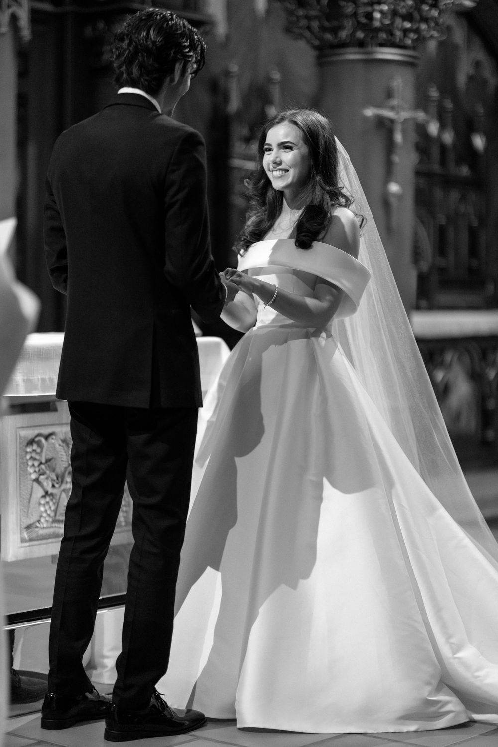 A bride in a flowing white ballgown with long veil stands with groom during wedding ceremony in elegant setting.