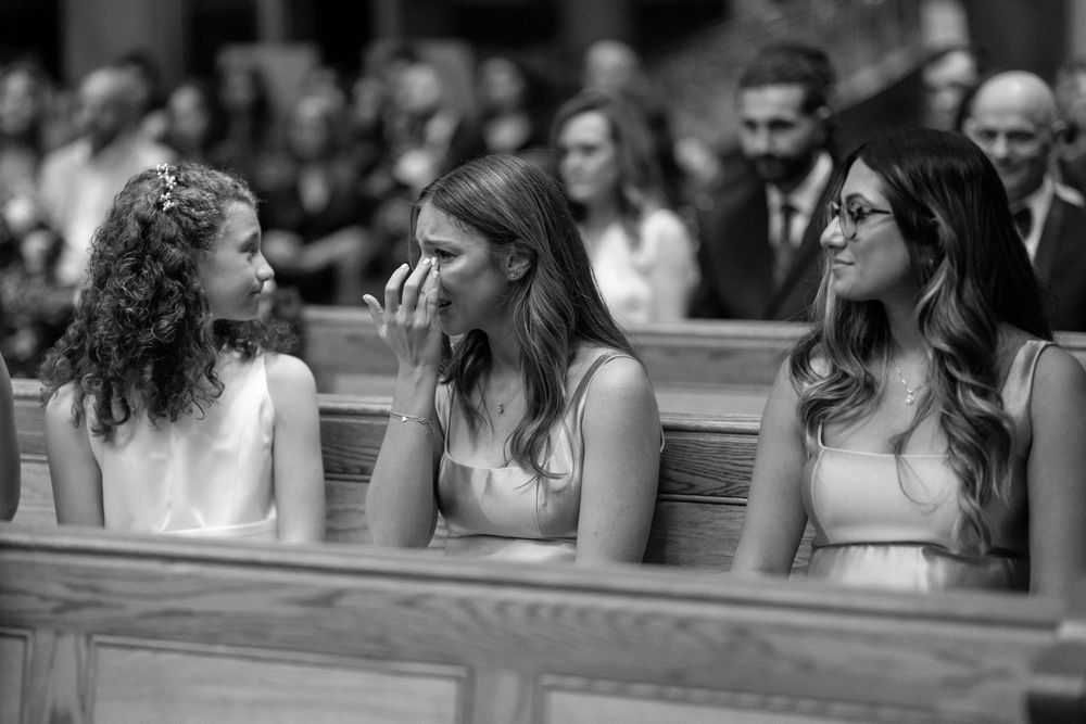 Three women in elegant dresses sit together in church pews during what appears to be a formal ceremony.