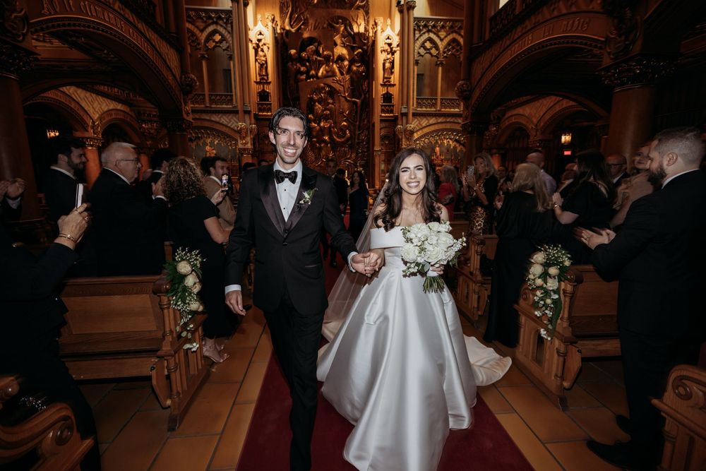 A couple walks down the aisle of an ornate church with gothic architecture and candlelit details during their wedding ceremony.