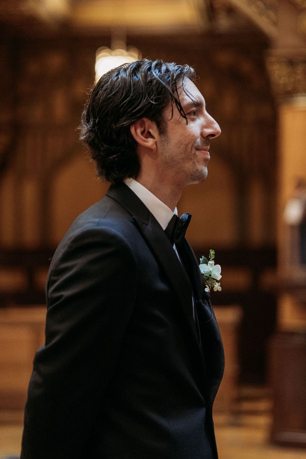 Profile view of a groom in a classic black tuxedo and bowtie with a floral boutonniere at a wedding ceremony.