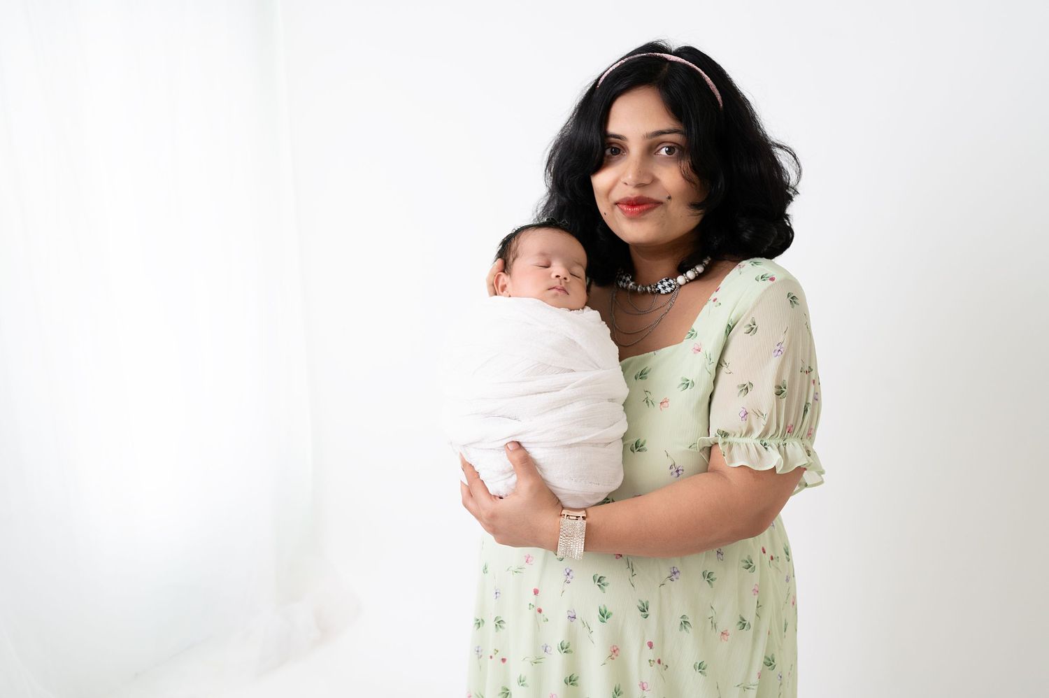 Mother in floral dress holding swaddled newborn against white background.