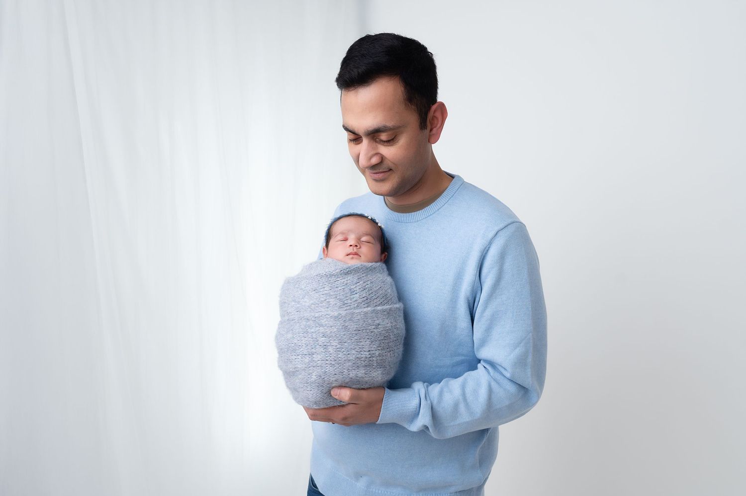 Parent holding swaddled newborn against white background while wearing light blue sweater.