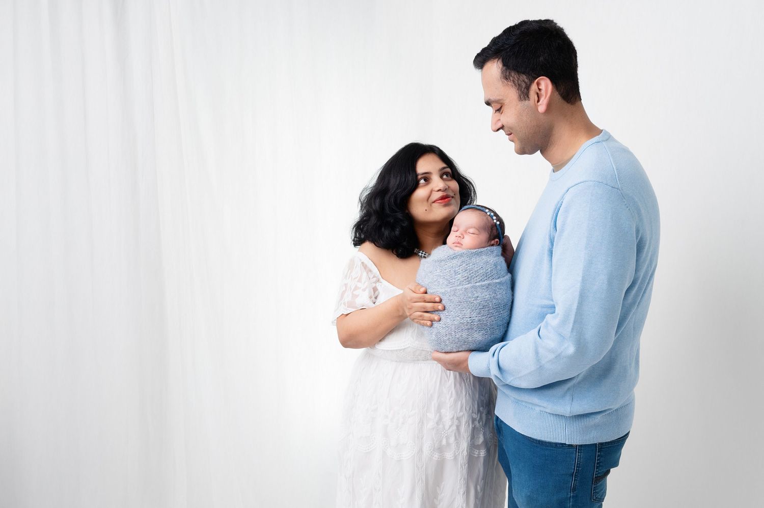 Happy parents in white and blue holding their swaddled newborn against white backdrop.