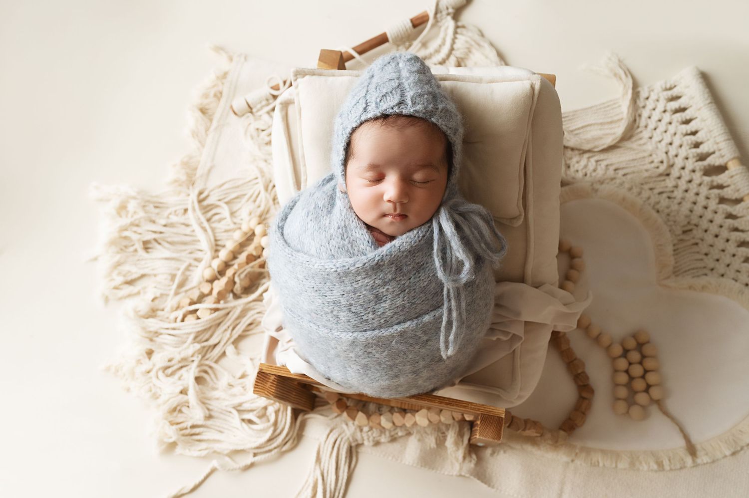 Newborn in gray knit wrap and bonnet sleeping on textured cream blanket.